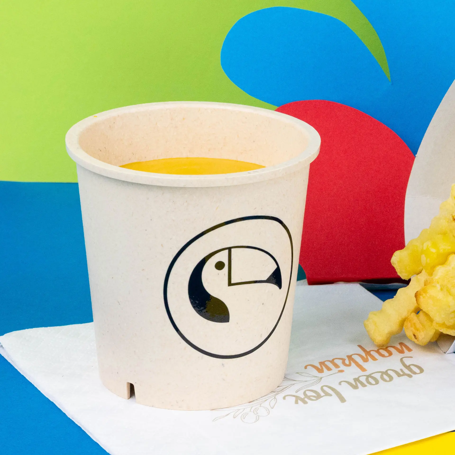 Round cream white reusable soup cup with a toucan logo, filled with yellow soup, placed on a napkin.