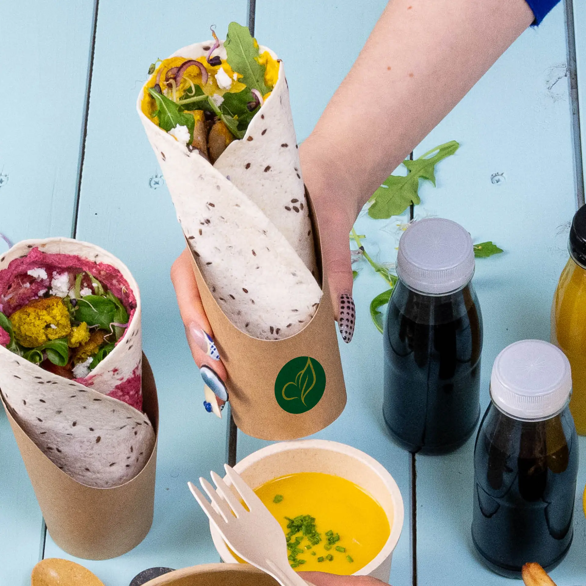 One brown custom printed wrap cup holding a filled wrap; another wrap cup beside it. Multiple bottles and a bowl with sauce are nearby.