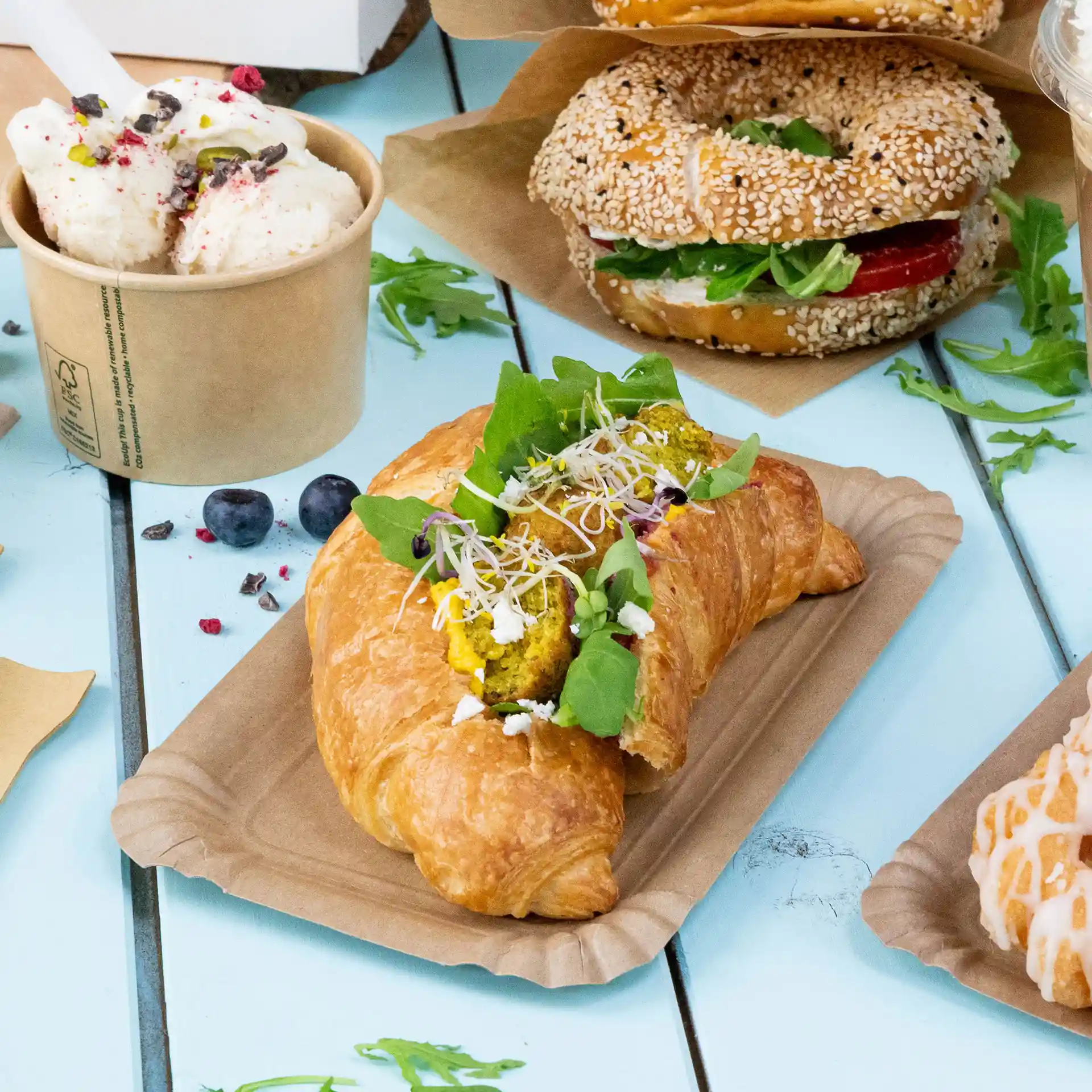 Croissant filled with green leaves, sprouts, and yellow filling on a brown paper plate, with other food items in the background.