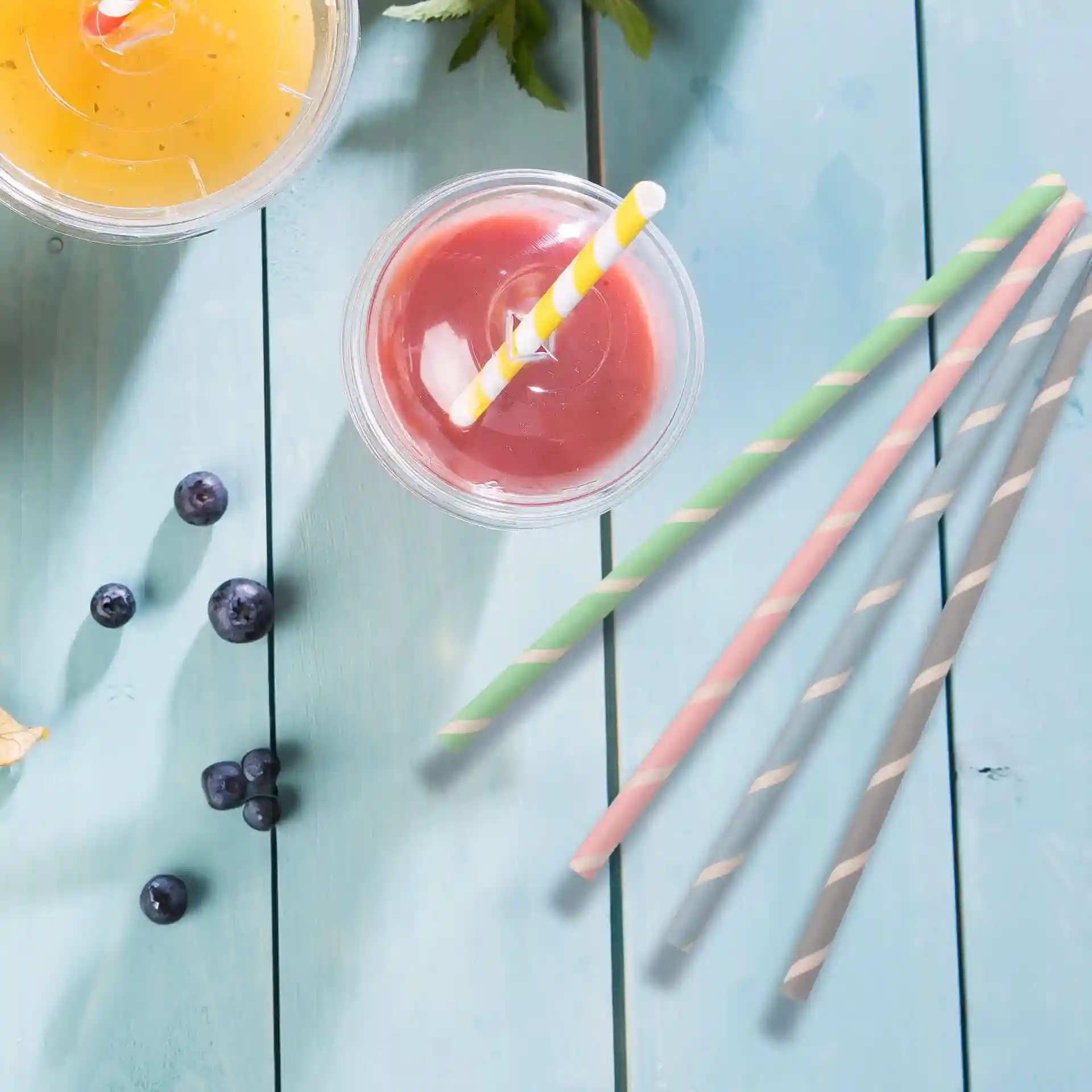 Two cups with drinks, one orange with a yellow straw and one red with a white and yellow striped straw. Nearby are several blueberries and four paper straws in green, pink, and gray.