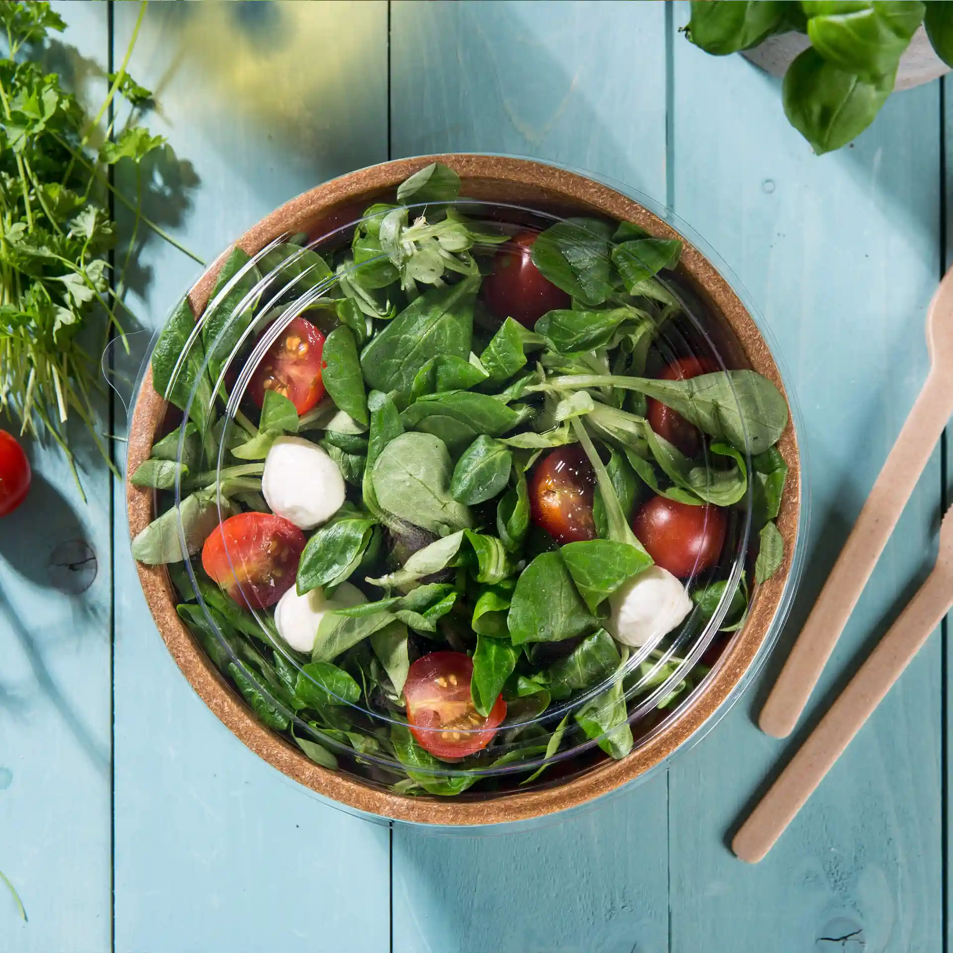 Clear round cup lid made of PLA, placed over a bowl of salad with tomatoes and spinach on a blue wooden surface.