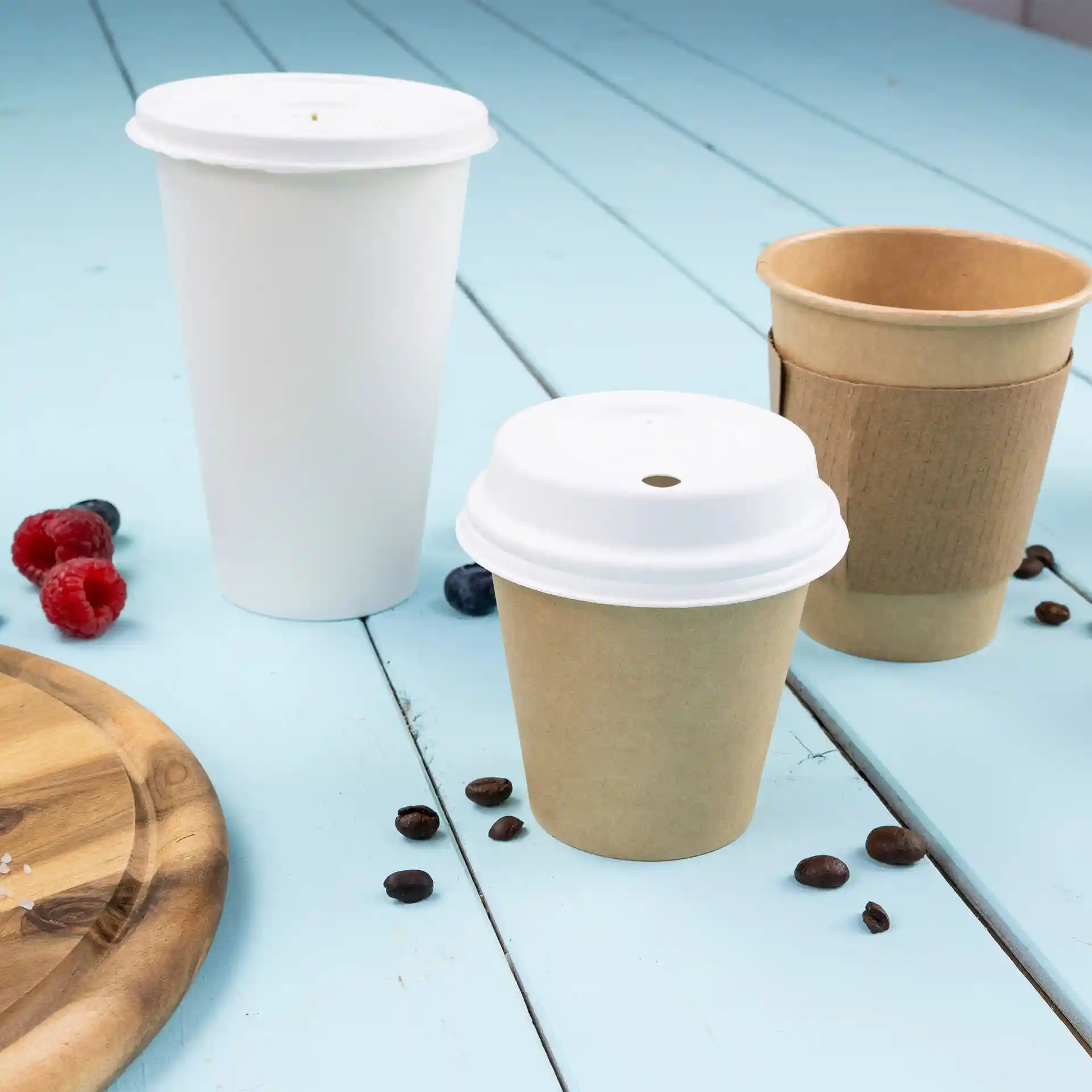 Image of three paper cups: one white, one brown with a white lid, and one brown without a lid, on a blue surface with coffee beans and berries.