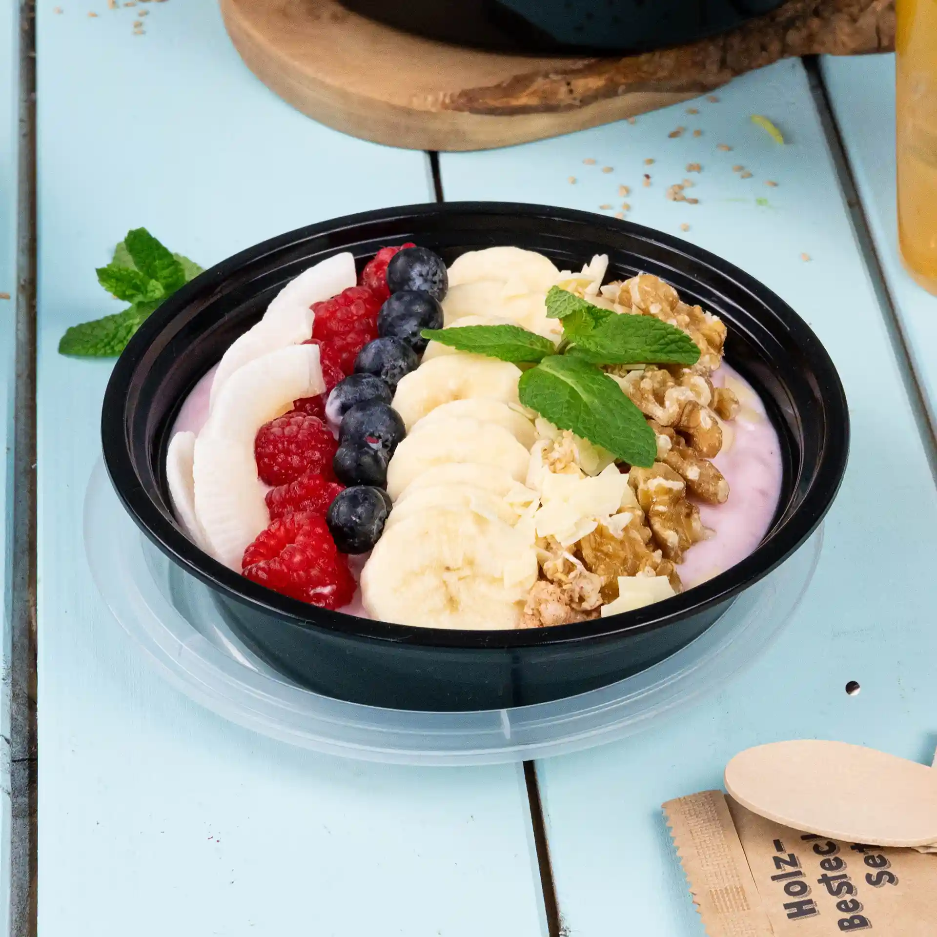 Bowl filled with yogurt, banana slices, raspberries, blueberries, granola, and mint leaves on a light blue surface.