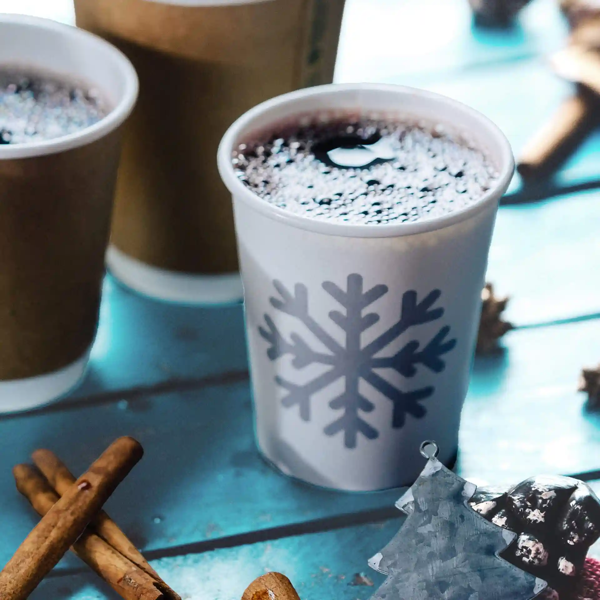 White paper cup with a snowflake design, filled with dark liquid, surrounded by cinnamon sticks and other cups.