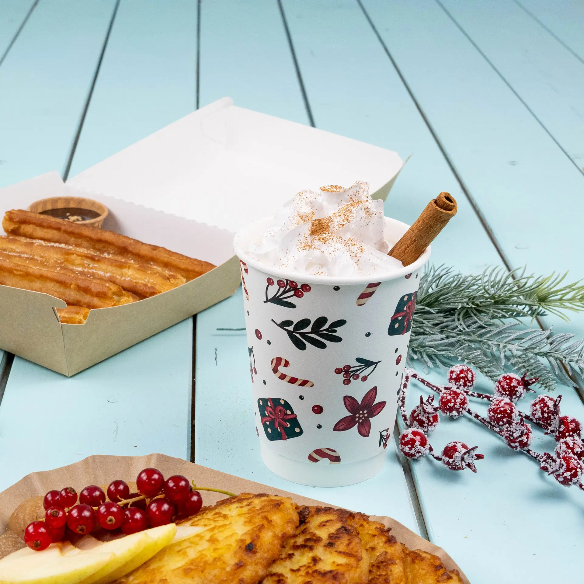 Double-wall coffee cup with winter design, filled with whipped cream and a cinnamon stick, next to churros and garnishes.