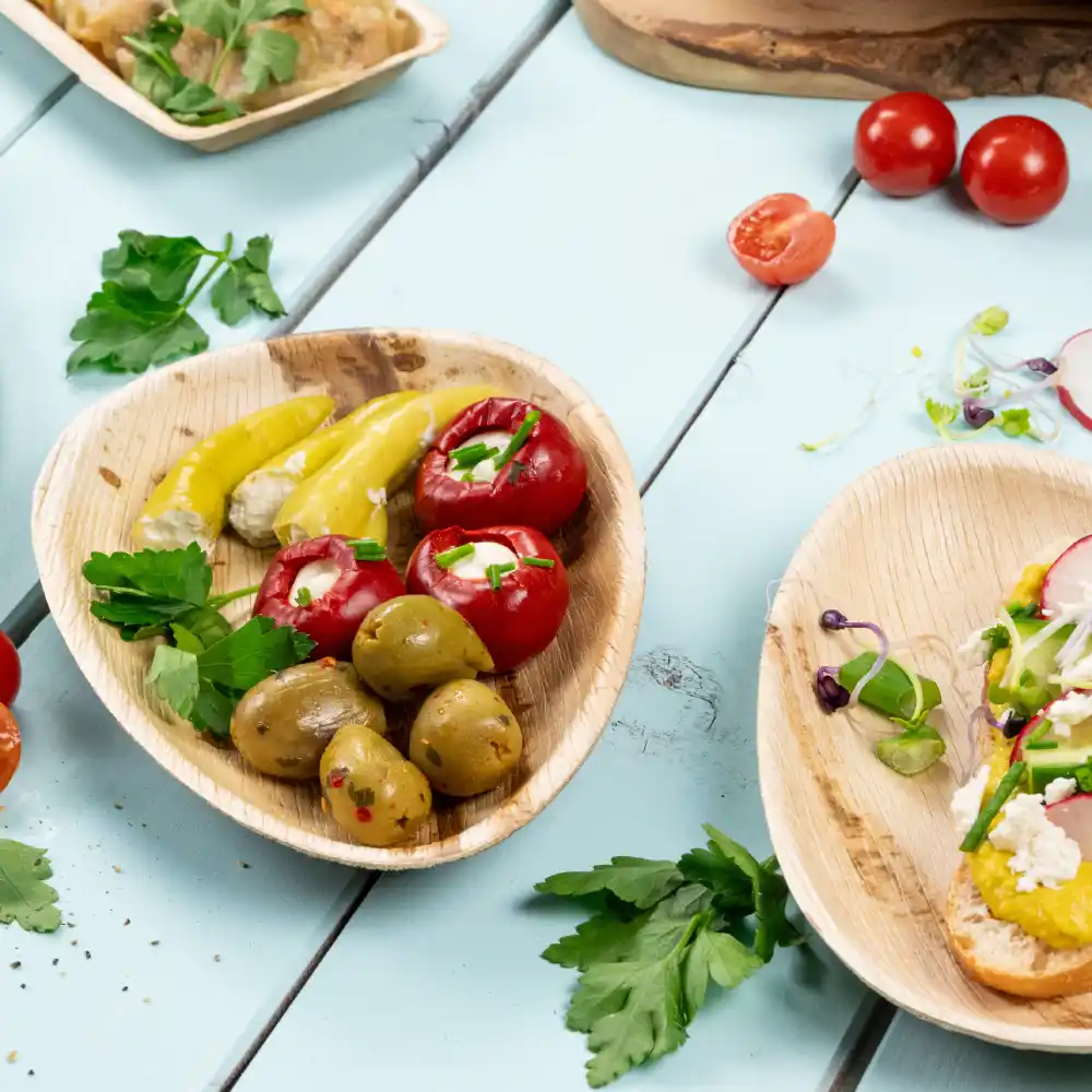 A wooden plate with stuffed red peppers, pickled yellow peppers, and green olives; fresh herbs scattered on a blue surface.