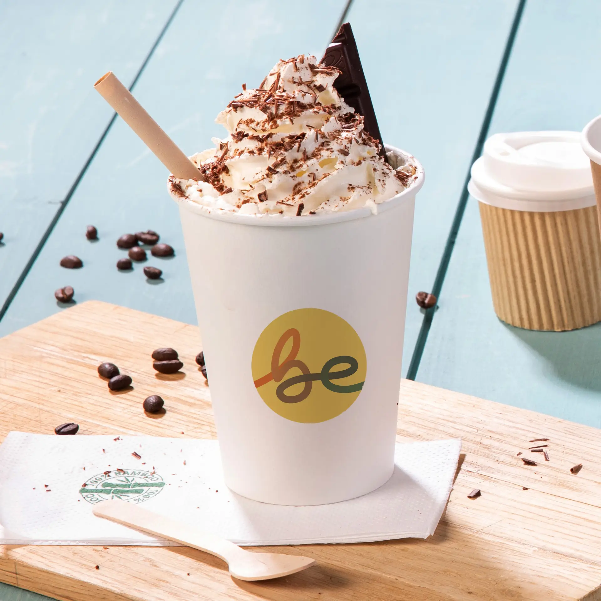 Cup of whipped cream topped with chocolate shavings and a chocolate square, with a printed logo and a wooden spoon on a serving board.