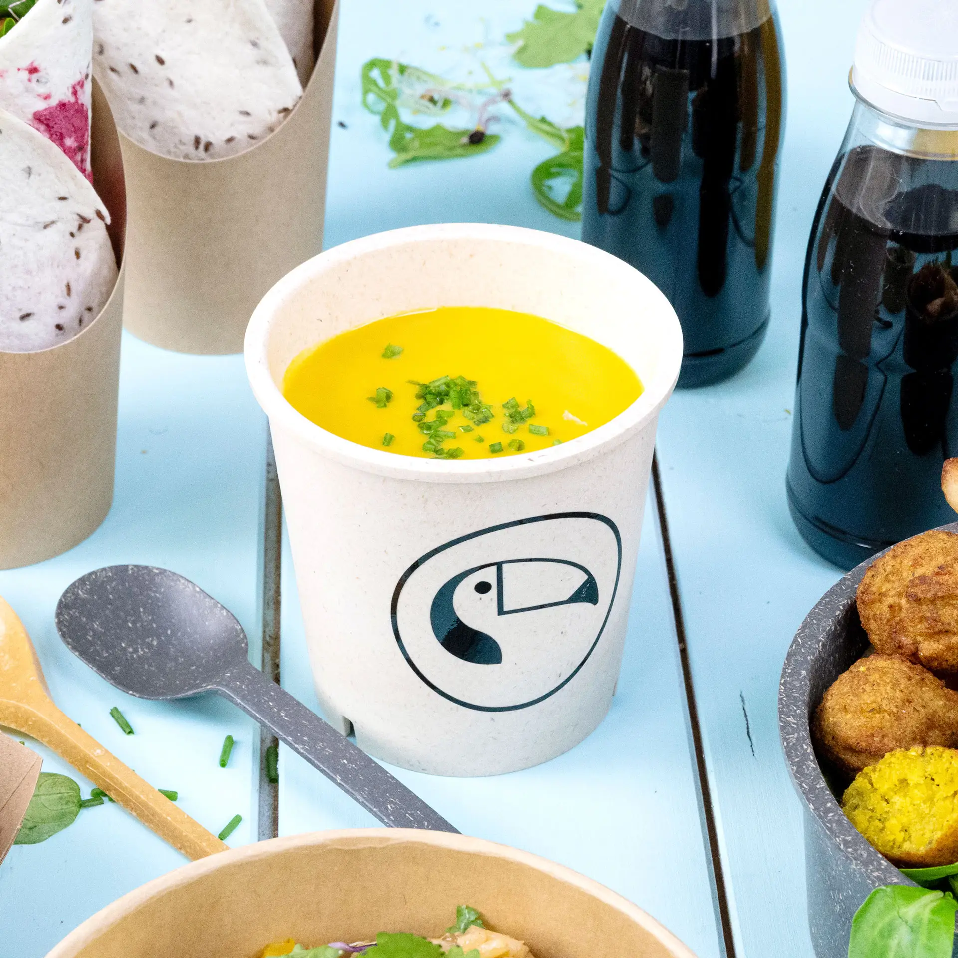 Round cream white soup cup with a logo, filled with yellow soup and topped with chopped chives, on a blue surface.