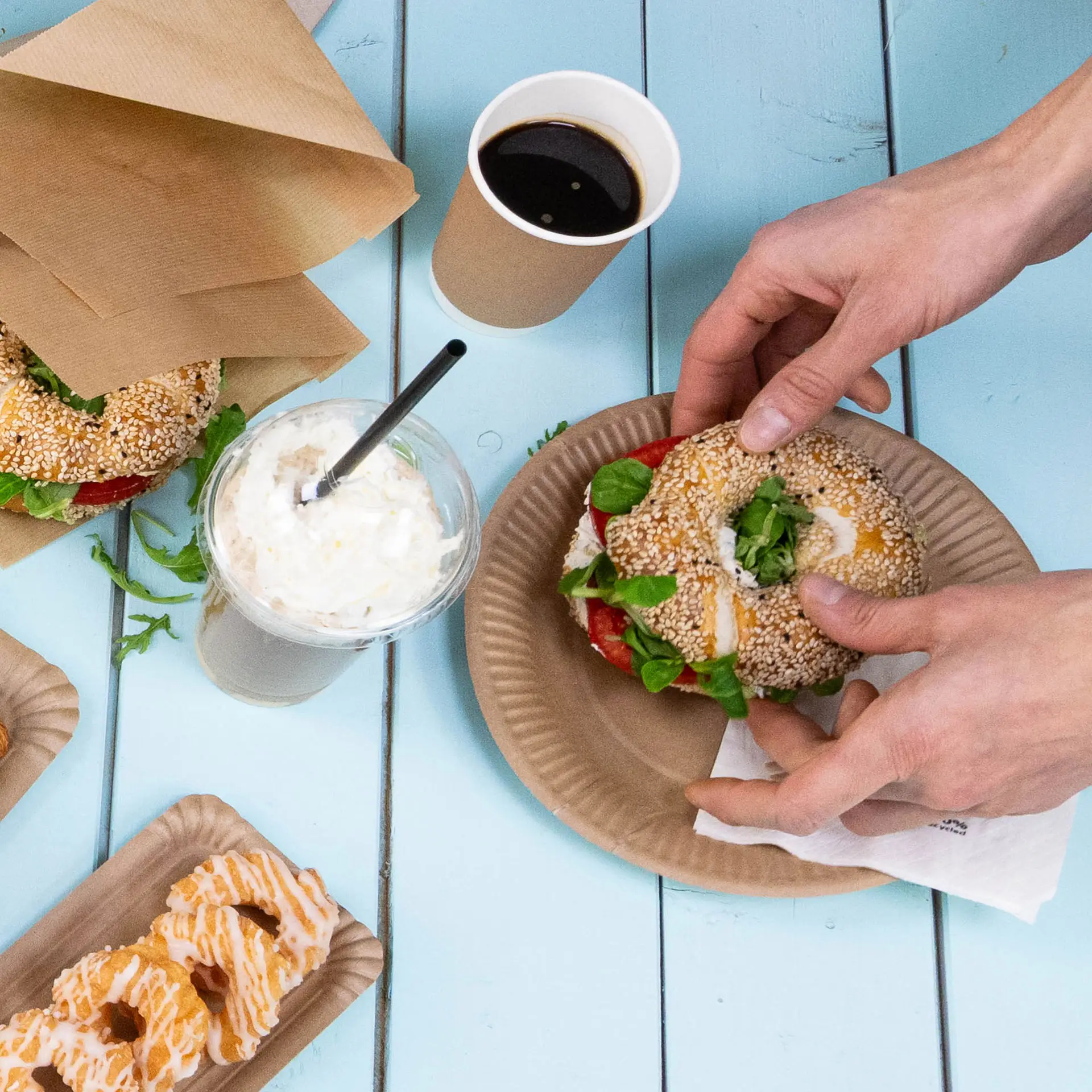 One hand holding a sesame seed bun with salad; several cardboard plates and drinks on a blue table, featuring multiple snacks.