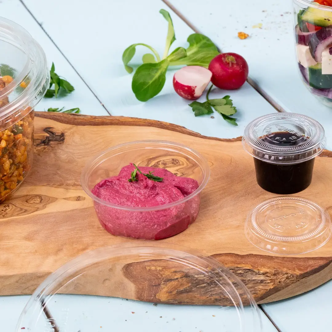 Clear plastic portion cup with pink dip, surrounded by a wooden board, salad ingredients, and another small cup.