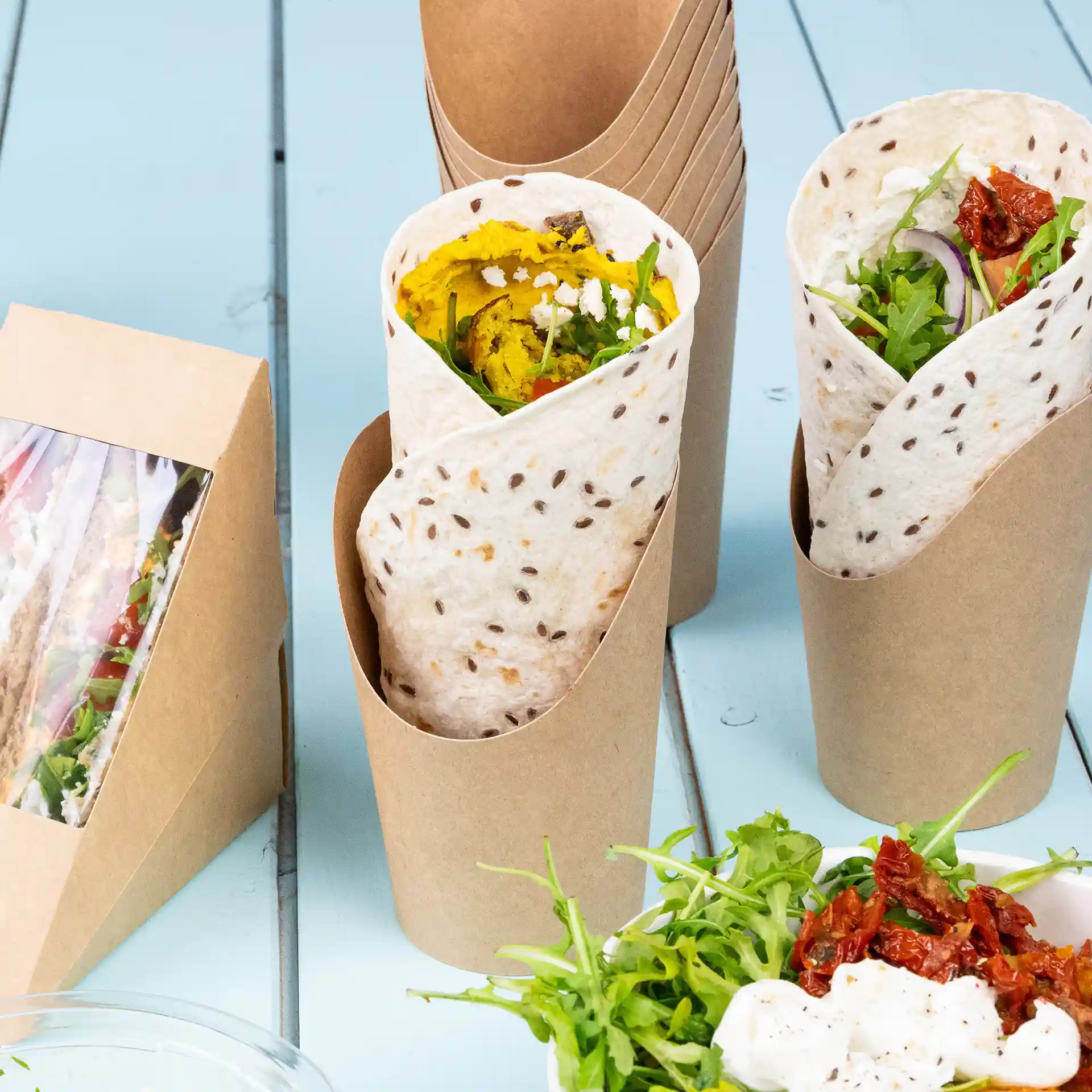 Image showing two wrap cups made of brown cardboard, one filled with food, and a side salad with greens and sun-dried tomatoes.