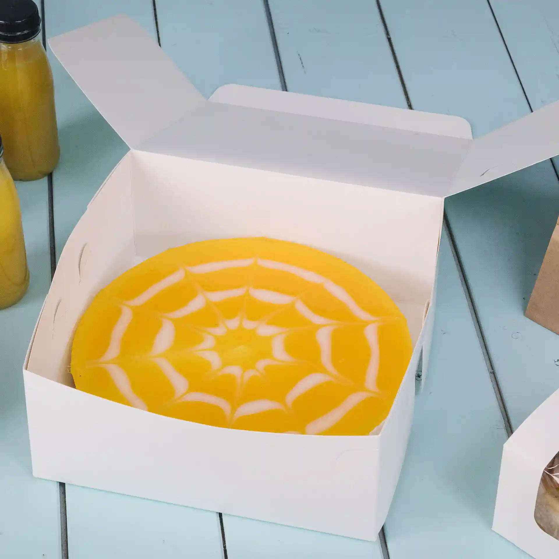 White cake box with a yellow cake featuring a spider web design inside, placed on a blue surface.