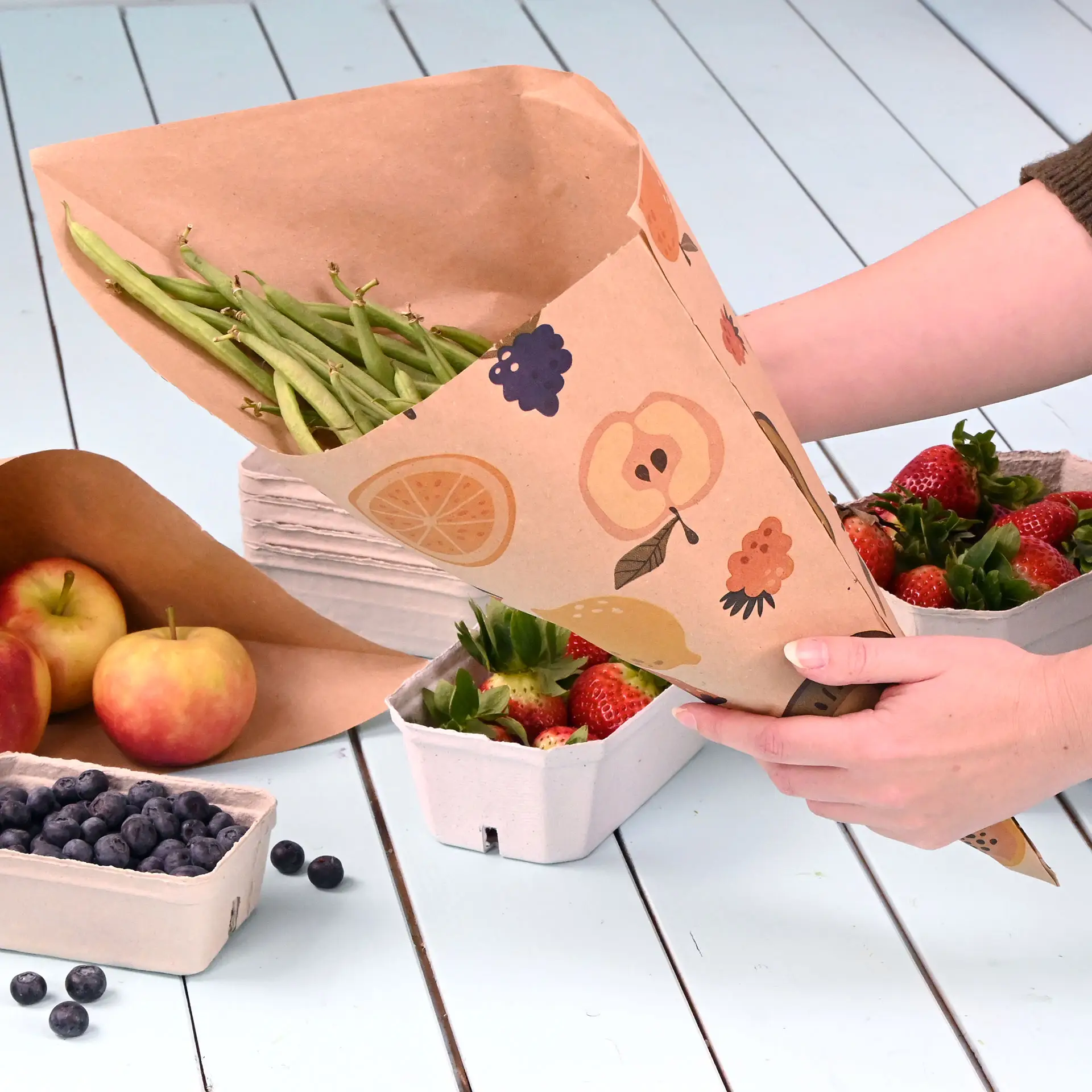 A hand holds a brown paper cone filled with green beans, surrounded by strawberries, apples, and blueberries on a light blue surface.