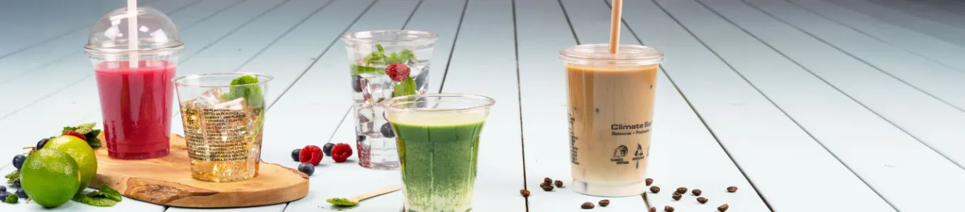 Assorted beverages displayed on a wooden surface, including iced coffee, smoothie, yogurt parfait, and juice, with orange slice nearby.