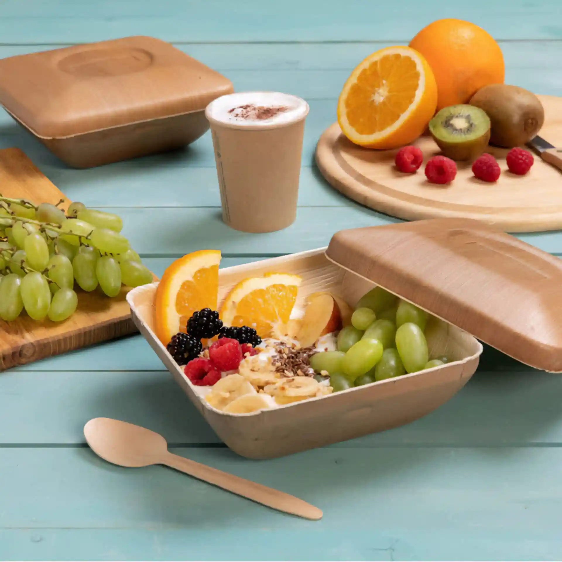 Square palm leaf bowl with lid containing fruit, spoon, and a cup on a blue wooden surface.