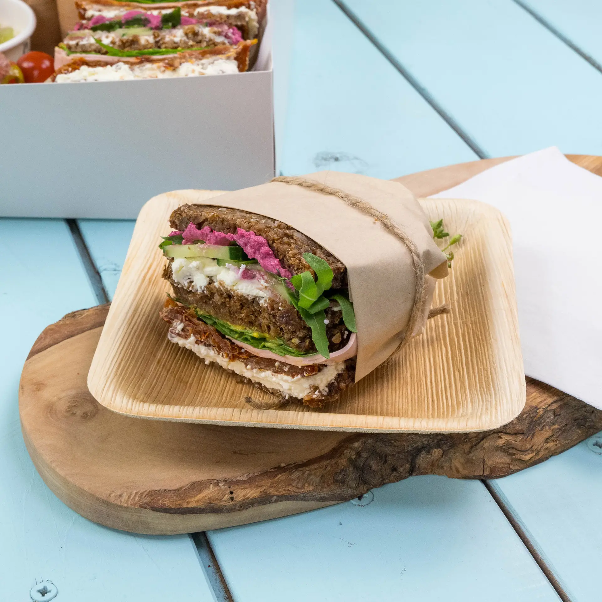 Brown wrapping paper with sandwich, featuring layers of greens, cream cheese, and pink spread on a wooden plate.