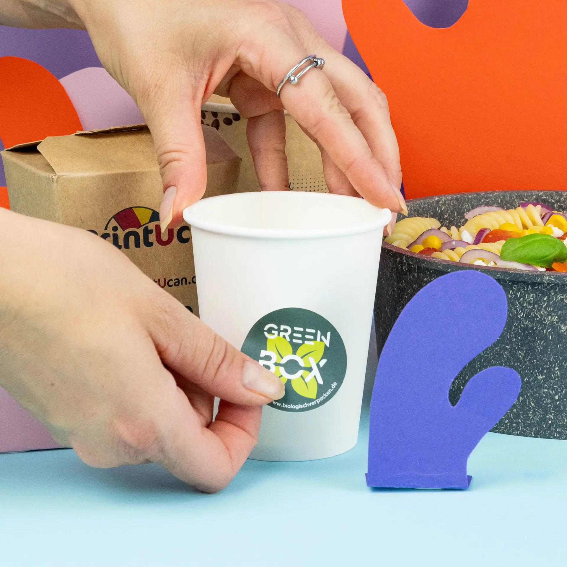 A hand is placing a round printed label on a white cup, with a purple oven mitt and a bowl of pasta in the background.