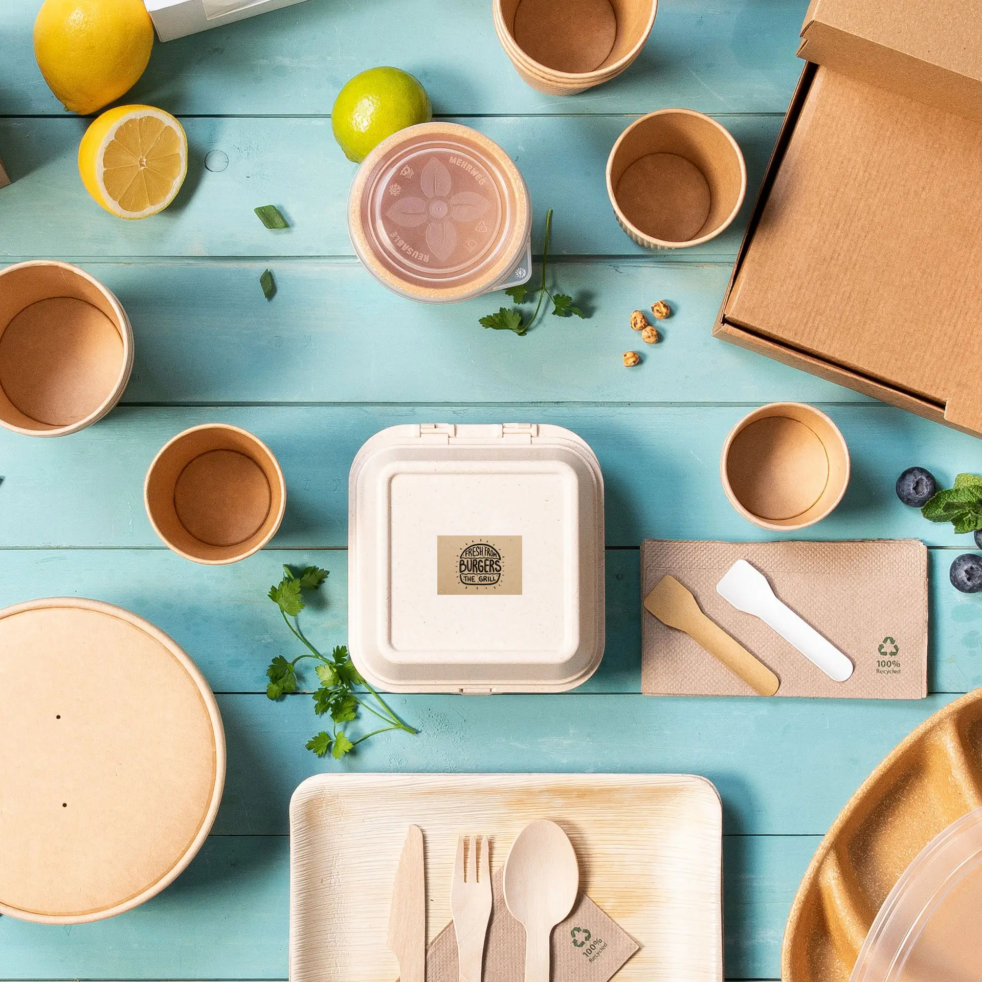 An assortment of eco-friendly packaging items, including bowls, cups, utensils, and a label, on a blue wooden surface.