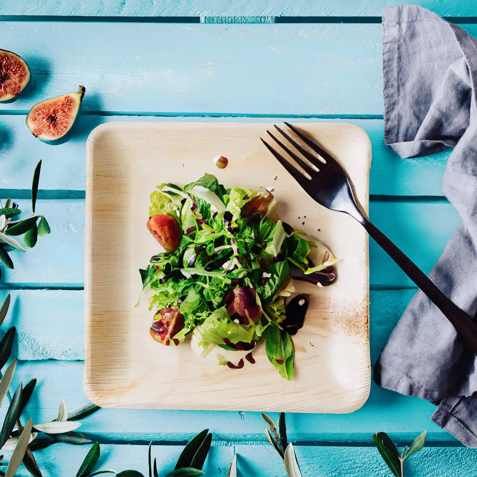 Square palm leaf plate with a salad topped with greens and figs, accompanied by a black fork on a blue wooden surface.