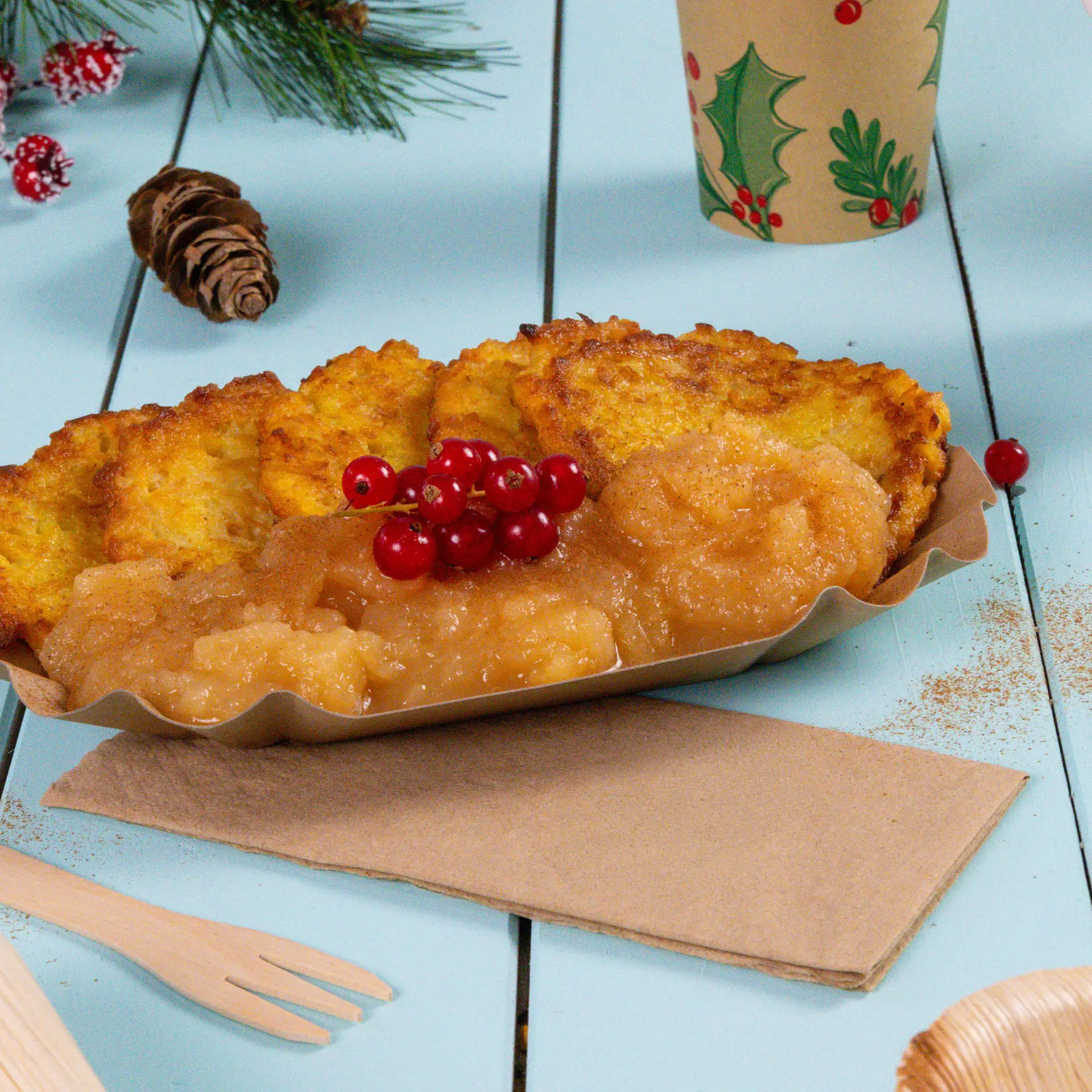 Plate with several golden-brown fritters, apple sauce, and red currants, on a light blue wooden surface with a napkin.