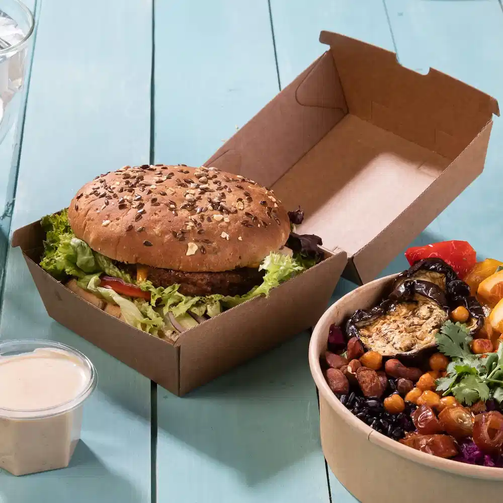 A sesame seed bun burger with lettuce in a brown takeaway box next to a bowl of mixed vegetables and legumes.