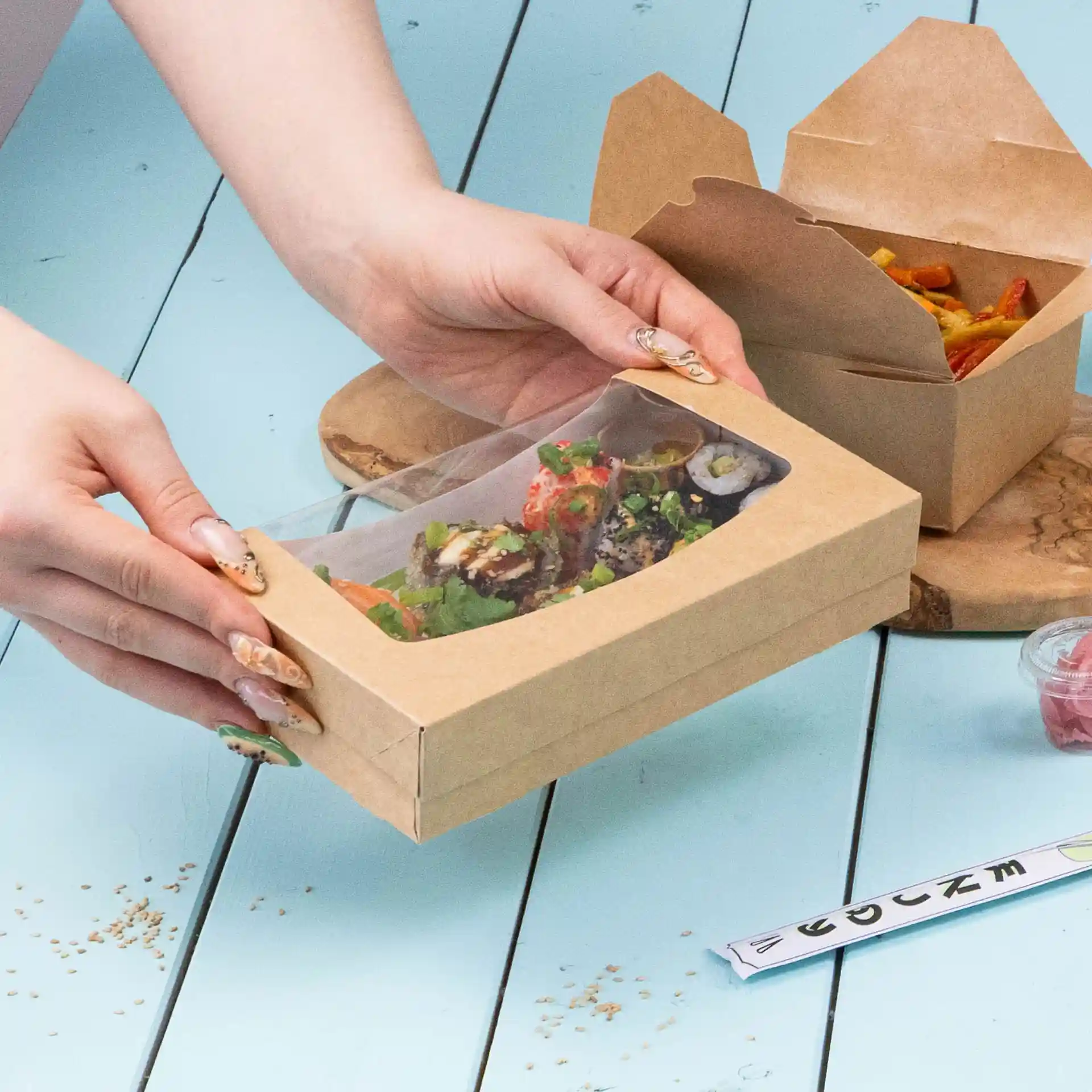 Cardboard window box holding sushi and salad, with a transparent top, on a blue surface.