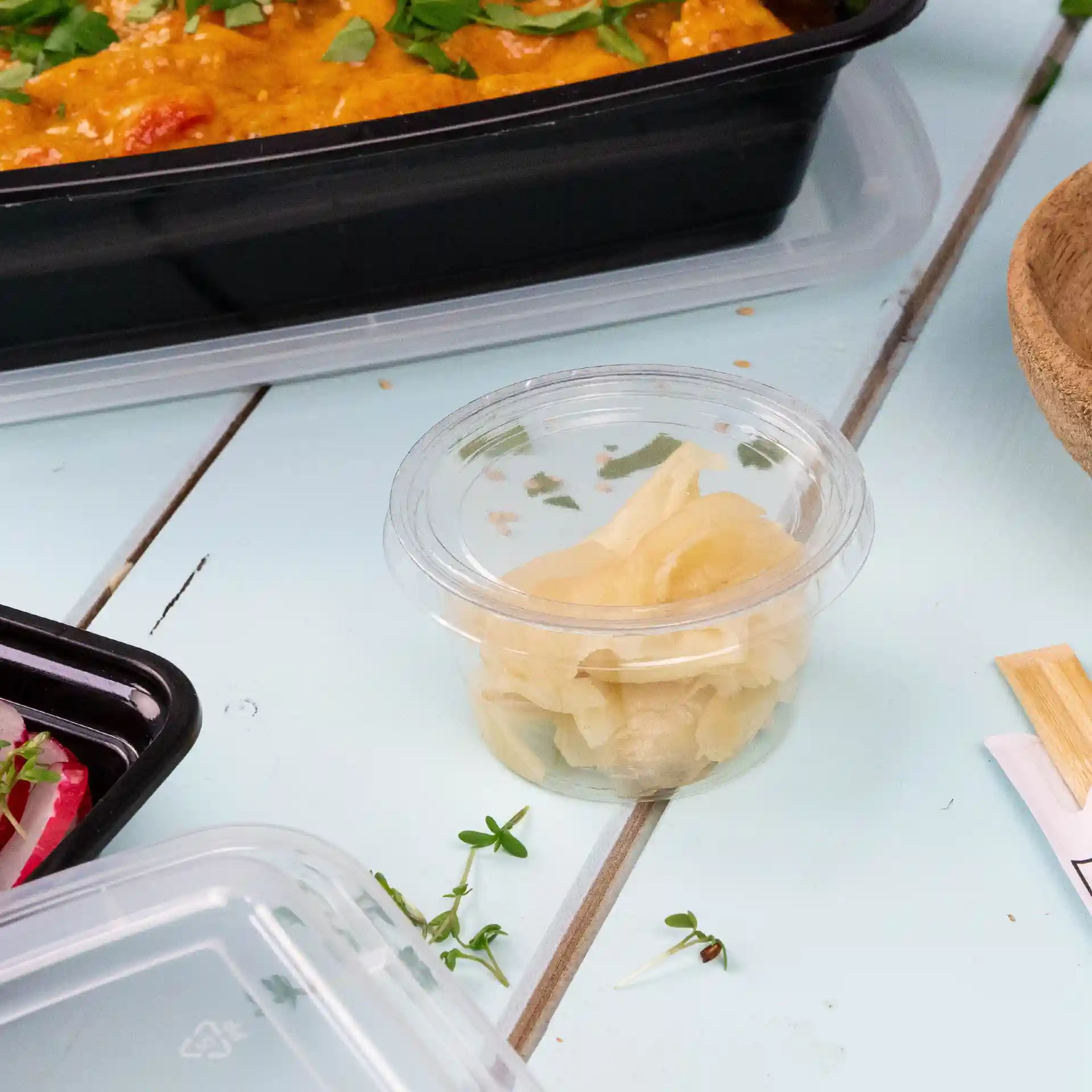 Clear PLA portion cup containing pickled ginger, placed on a light blue surface with other food items nearby.