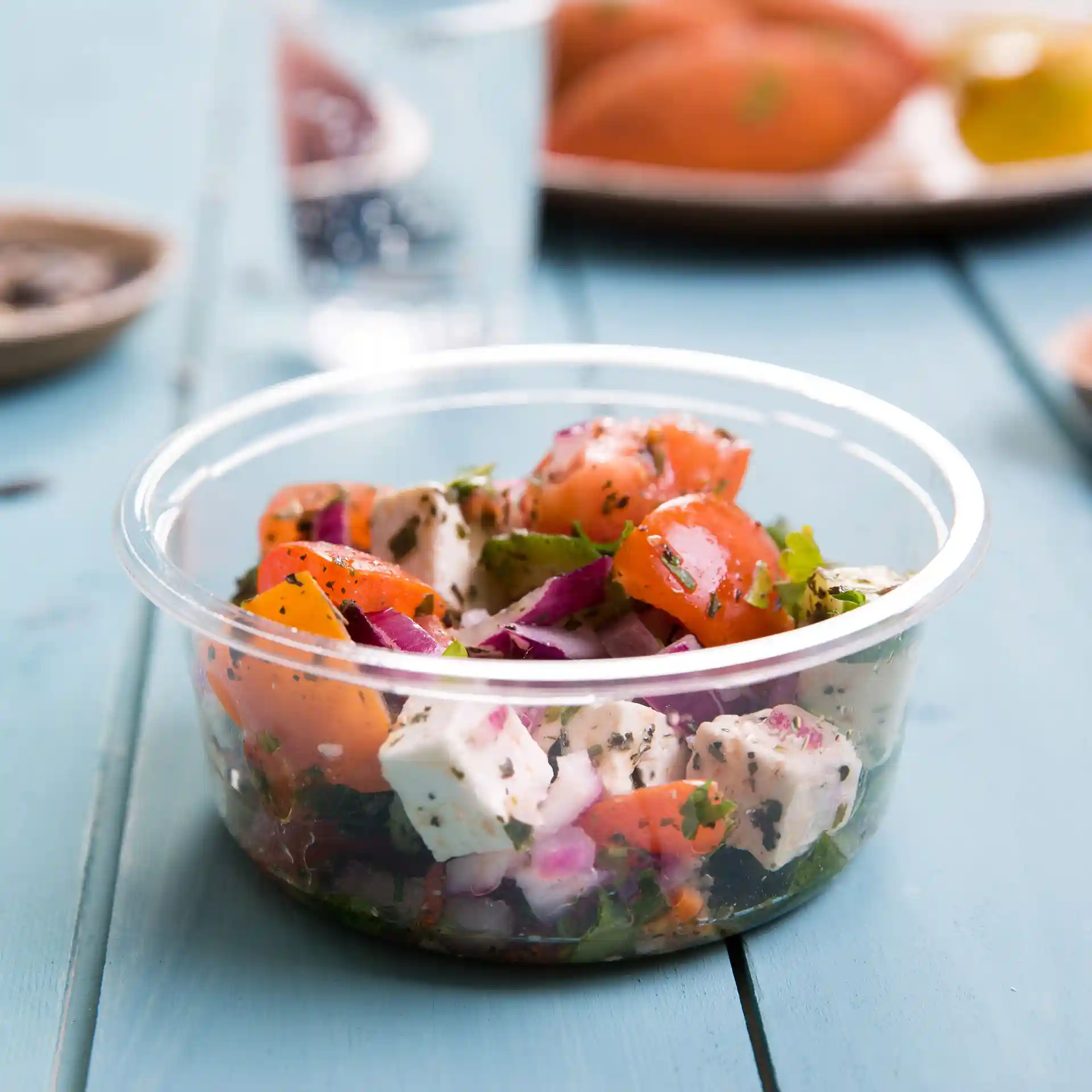 Transparent round deli container with a salad mix of tomatoes, onions, and herbs, placed on a blue wooden surface.