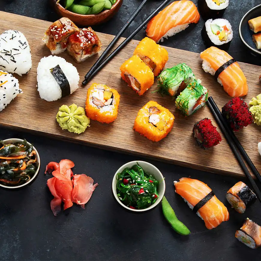 A wooden platter with various sushi types, including rolls, sashimi, and nigiri. Accompanied by wasabi, pickled ginger, and small bowls of seaweed salad.