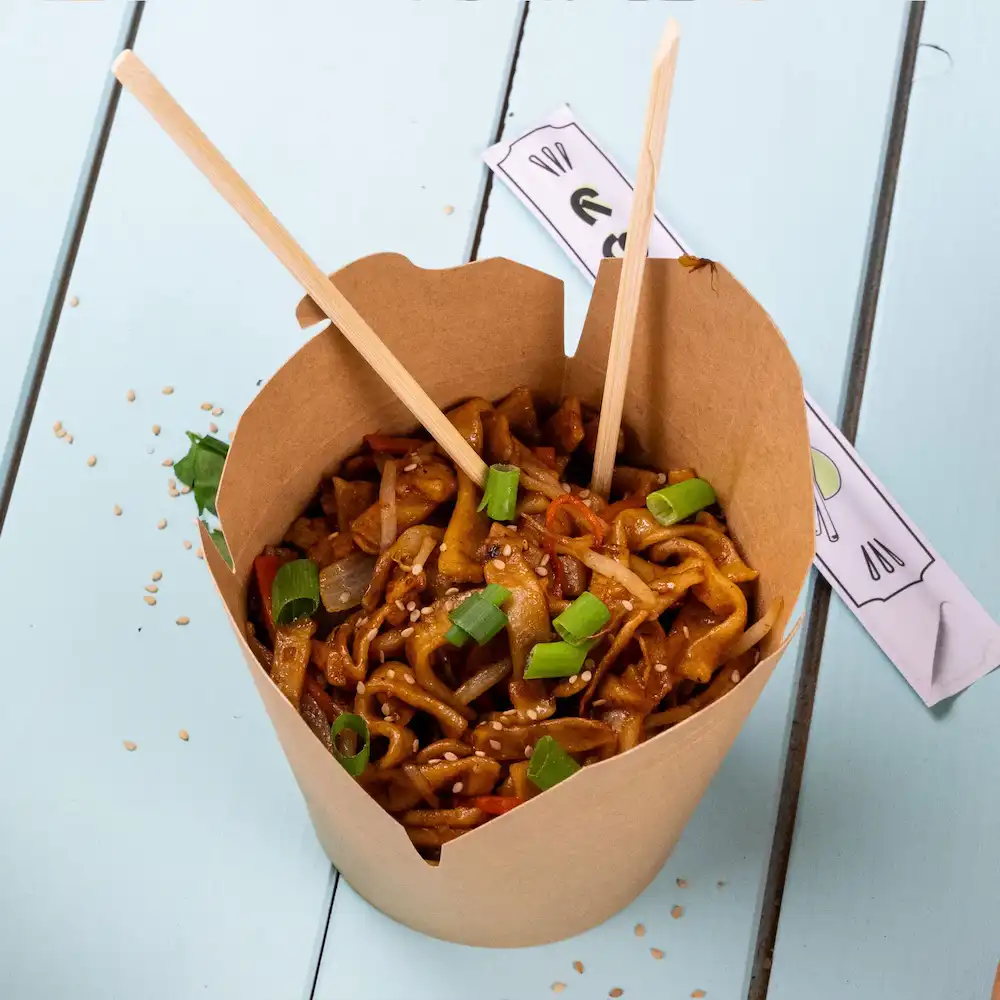 Box of stir-fried noodles with vegetables, topped with green onions and sesame seeds, served with chopsticks.