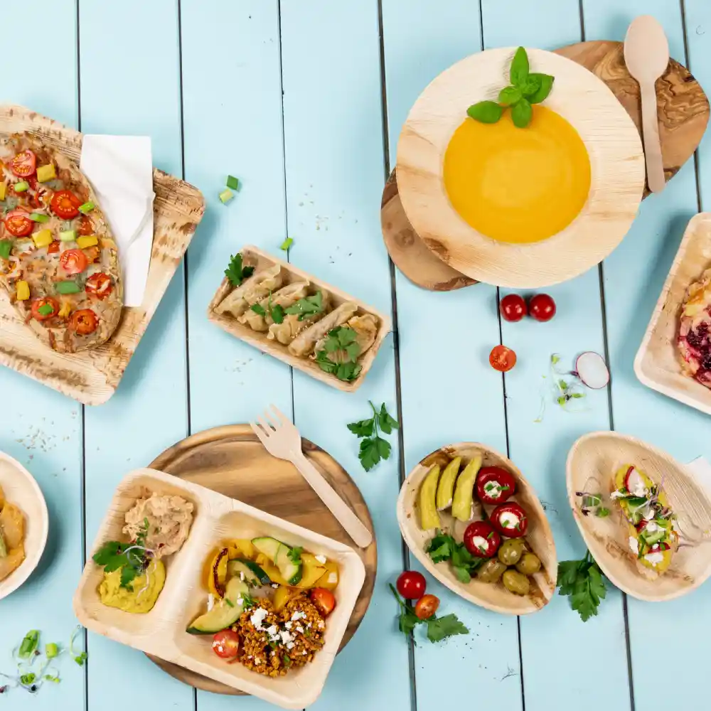 A variety of wooden disposable plates with colorful dishes, including salads, dips, and hummus, arranged on a blue wooden surface.