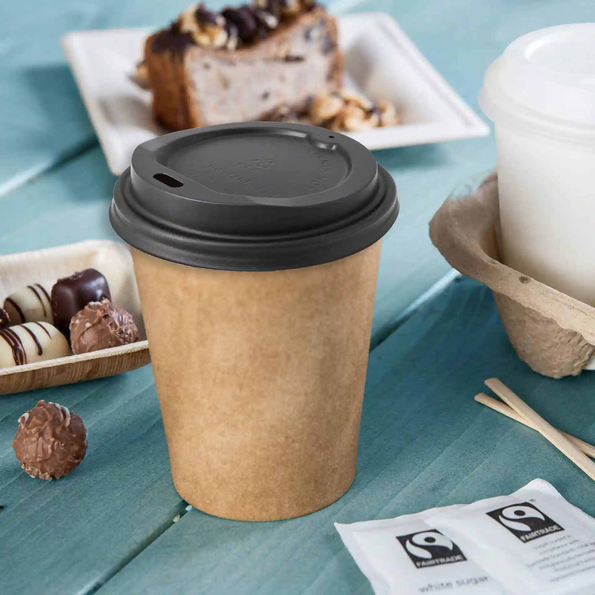 Black CPLA cup lid with a drinking hole, placed next to a brown paper cup on a blue wooden surface.
