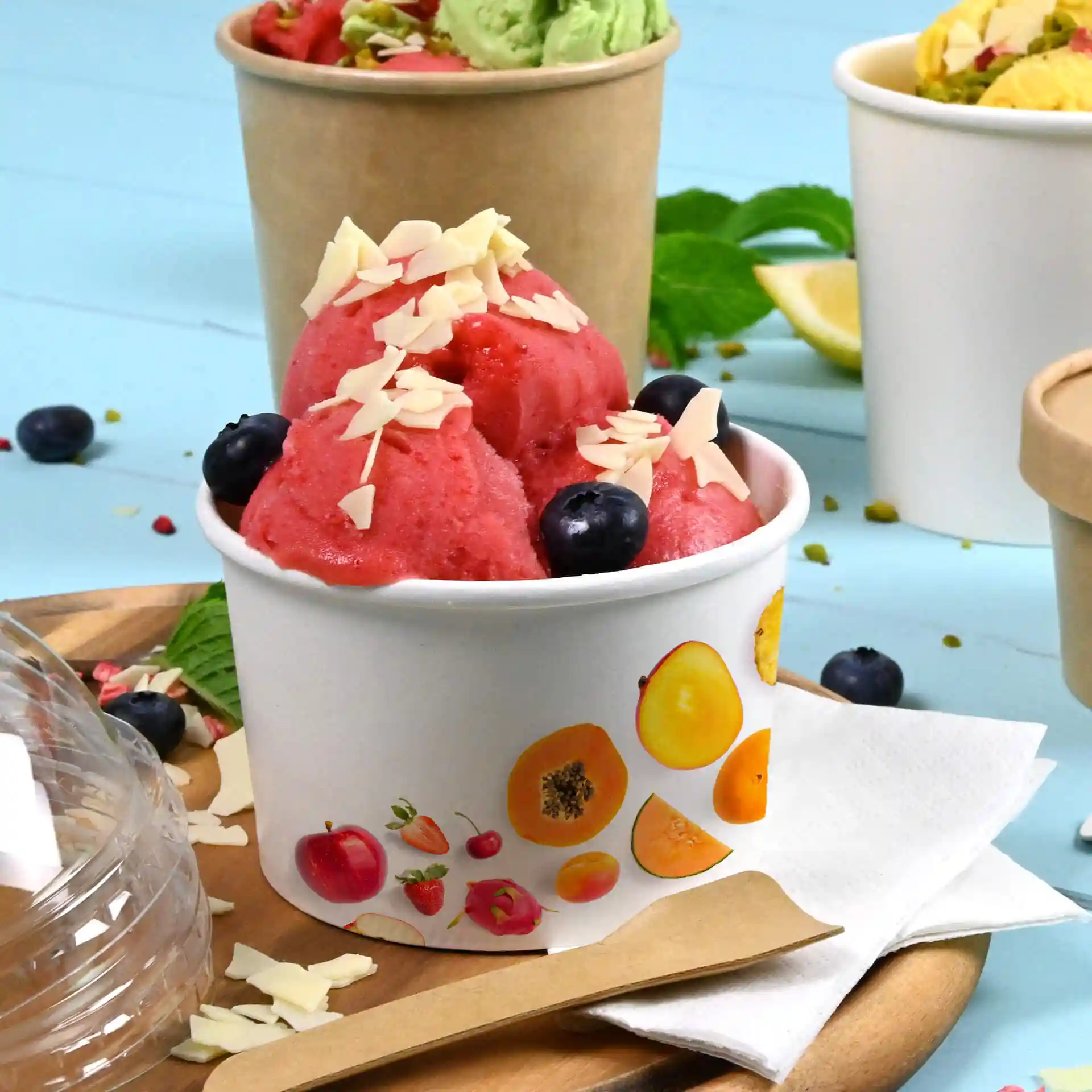Round white cardboard ice cream cup filled with pink sorbet, topped with almond flakes and blueberries, on a wooden tray.