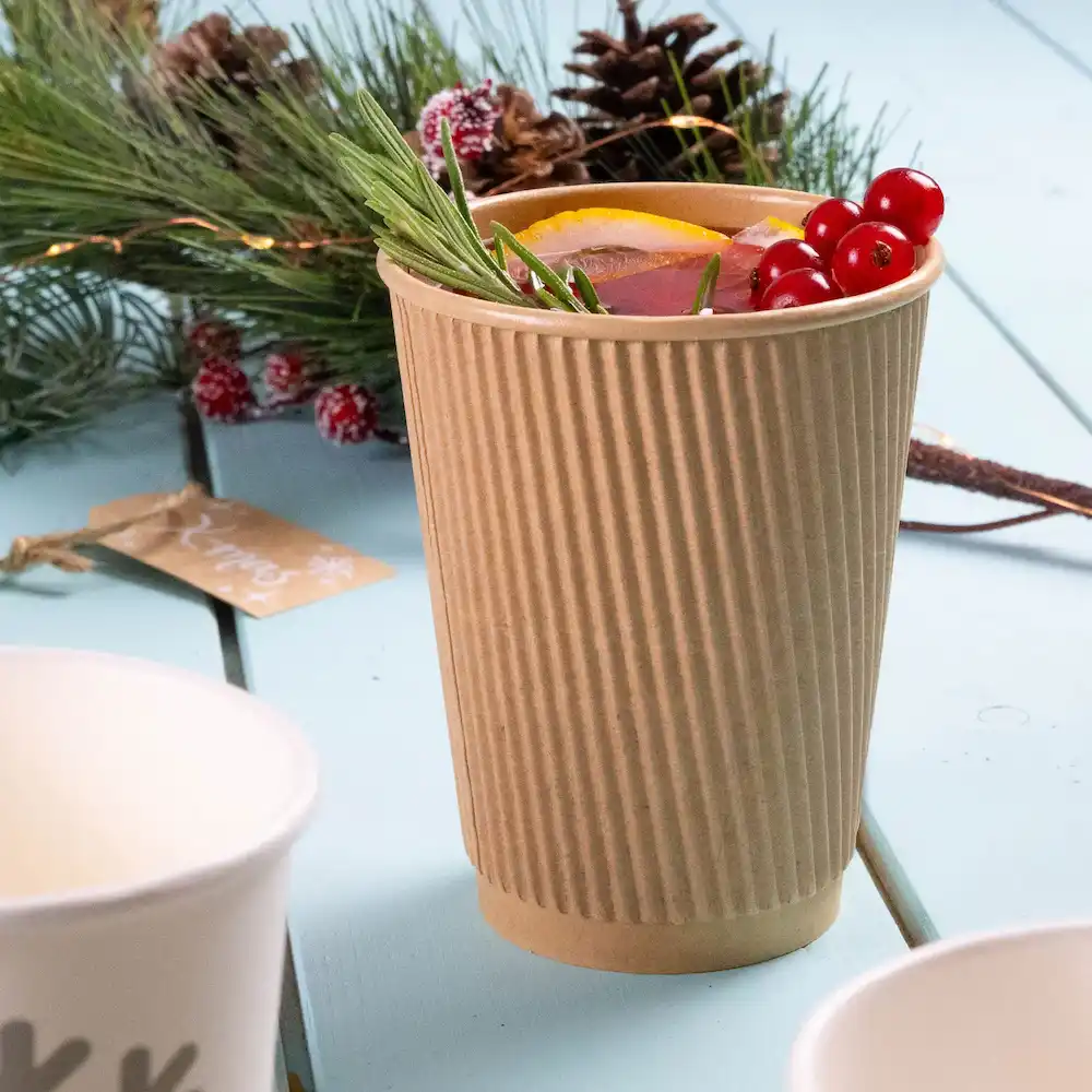 Corrugated brown paper cup filled with a drink, garnished with orange slices, rosemary, and red berries, on a festive table.