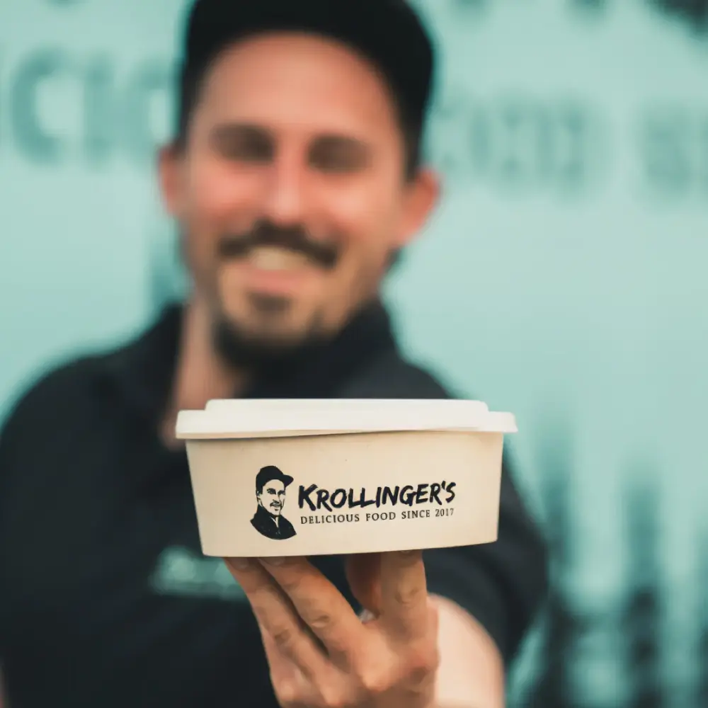 Man holding a takeout container labeled "Krollinger's Delicious Food Since 2017" with a smiling background.