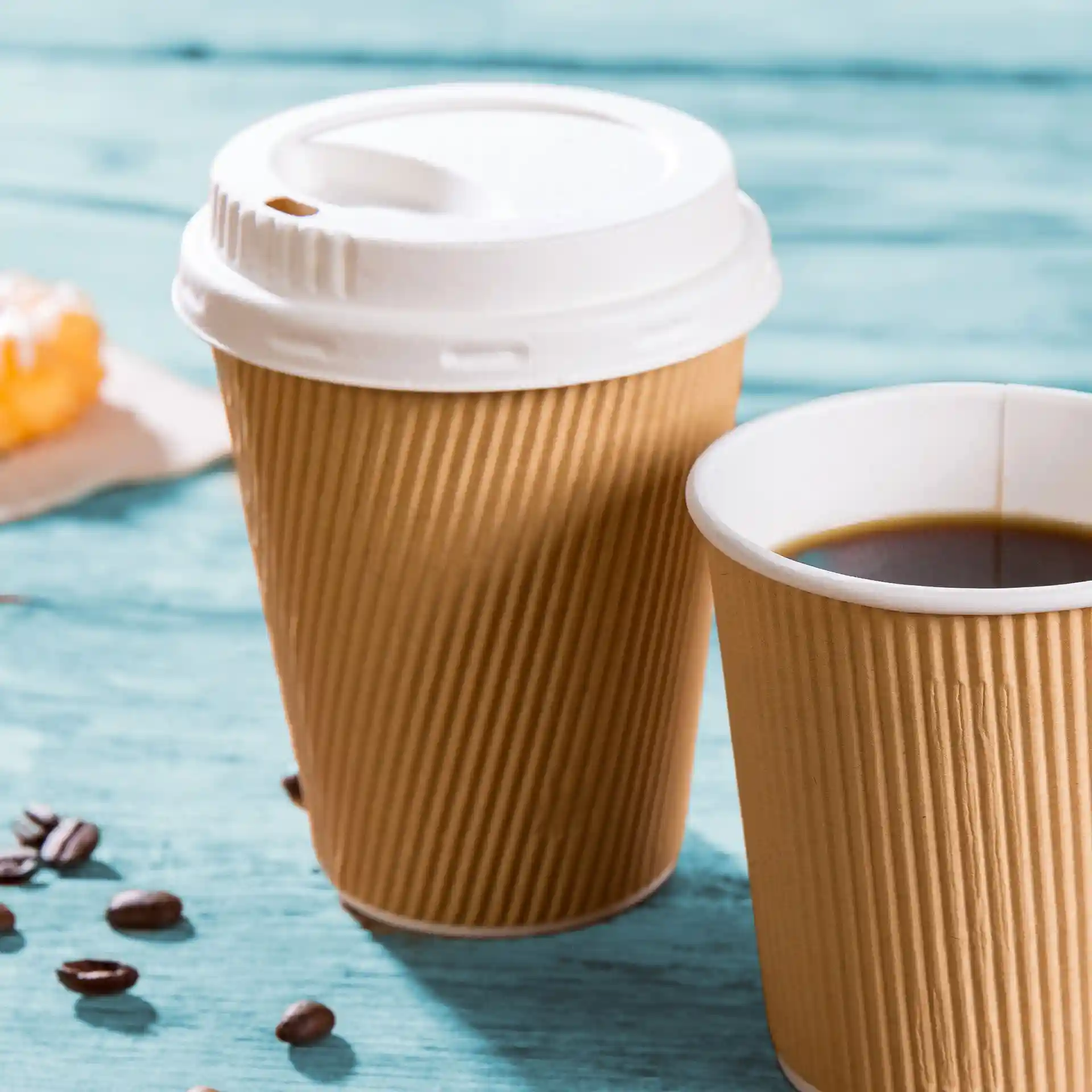 Two brown ripple coffee cups, one with a white lid, filled with coffee, on a blue wooden surface.