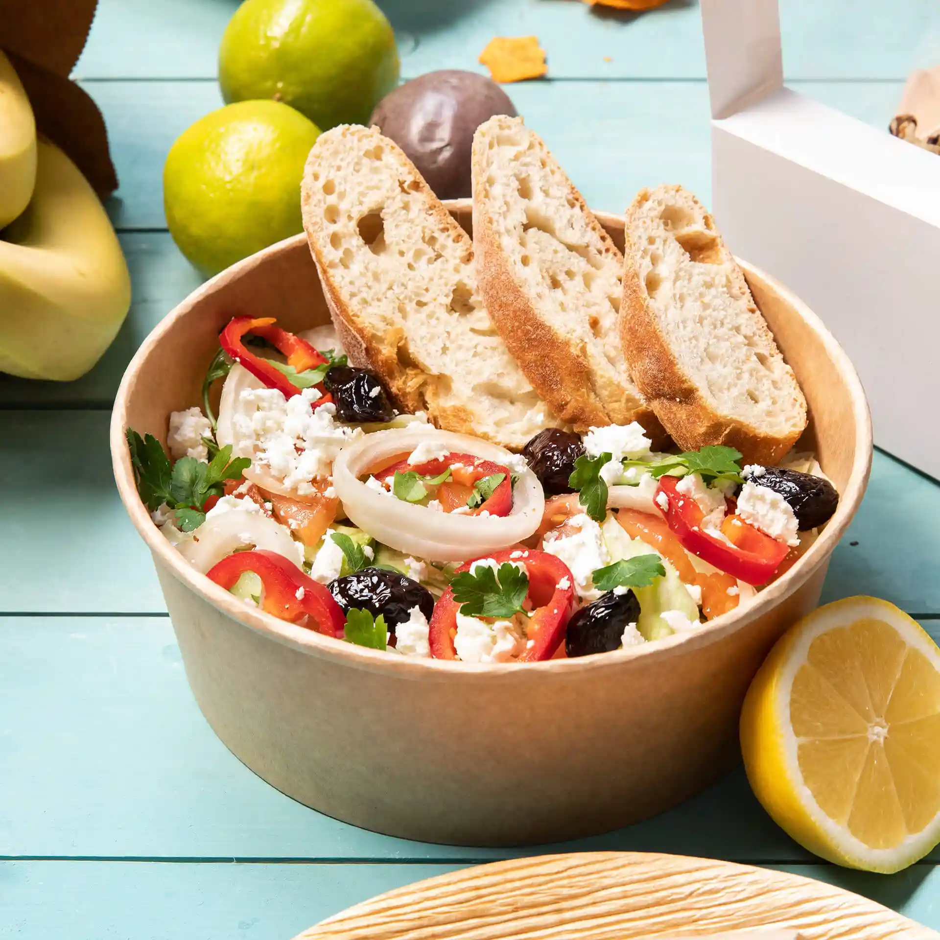 Round brown cardboard bowl filled with salad, topped with sliced bread, and surrounded by fruits on a blue surface.