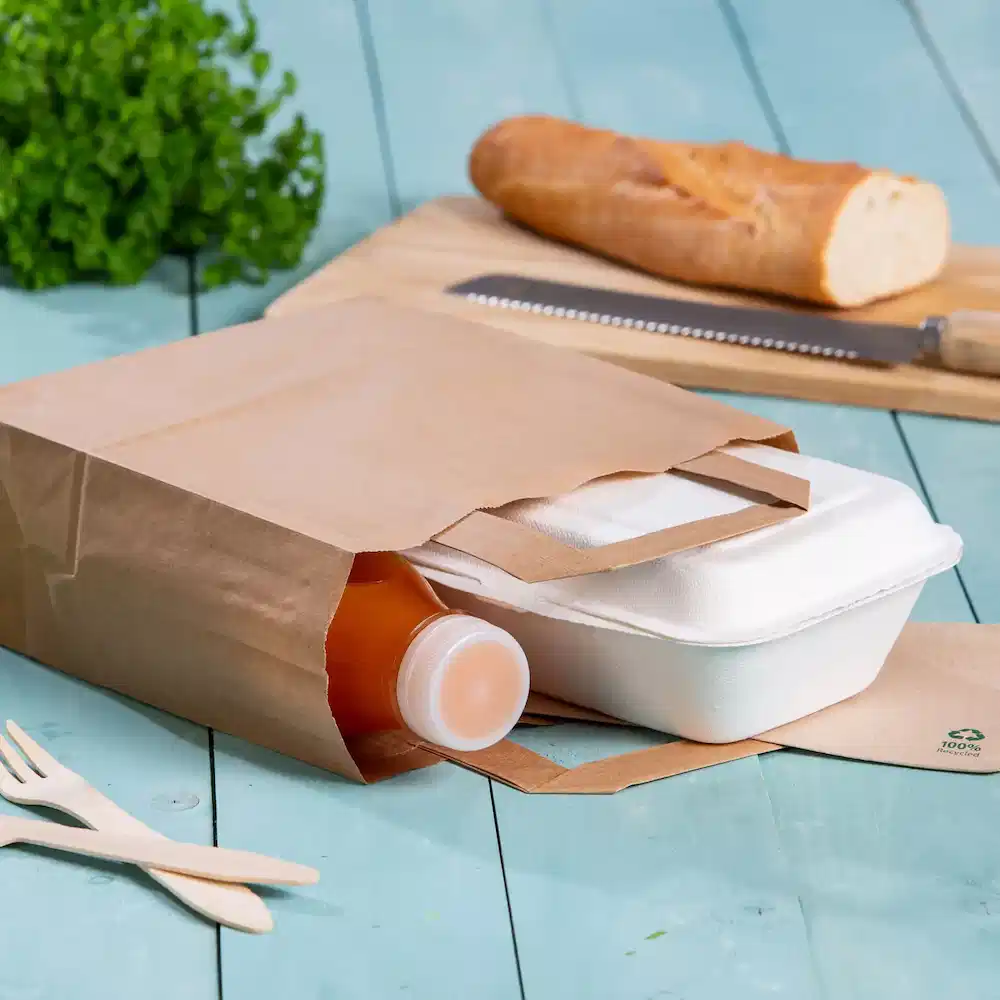 Brown paper bag containing a beverage bottle and a takeout food container on a blue wooden surface.