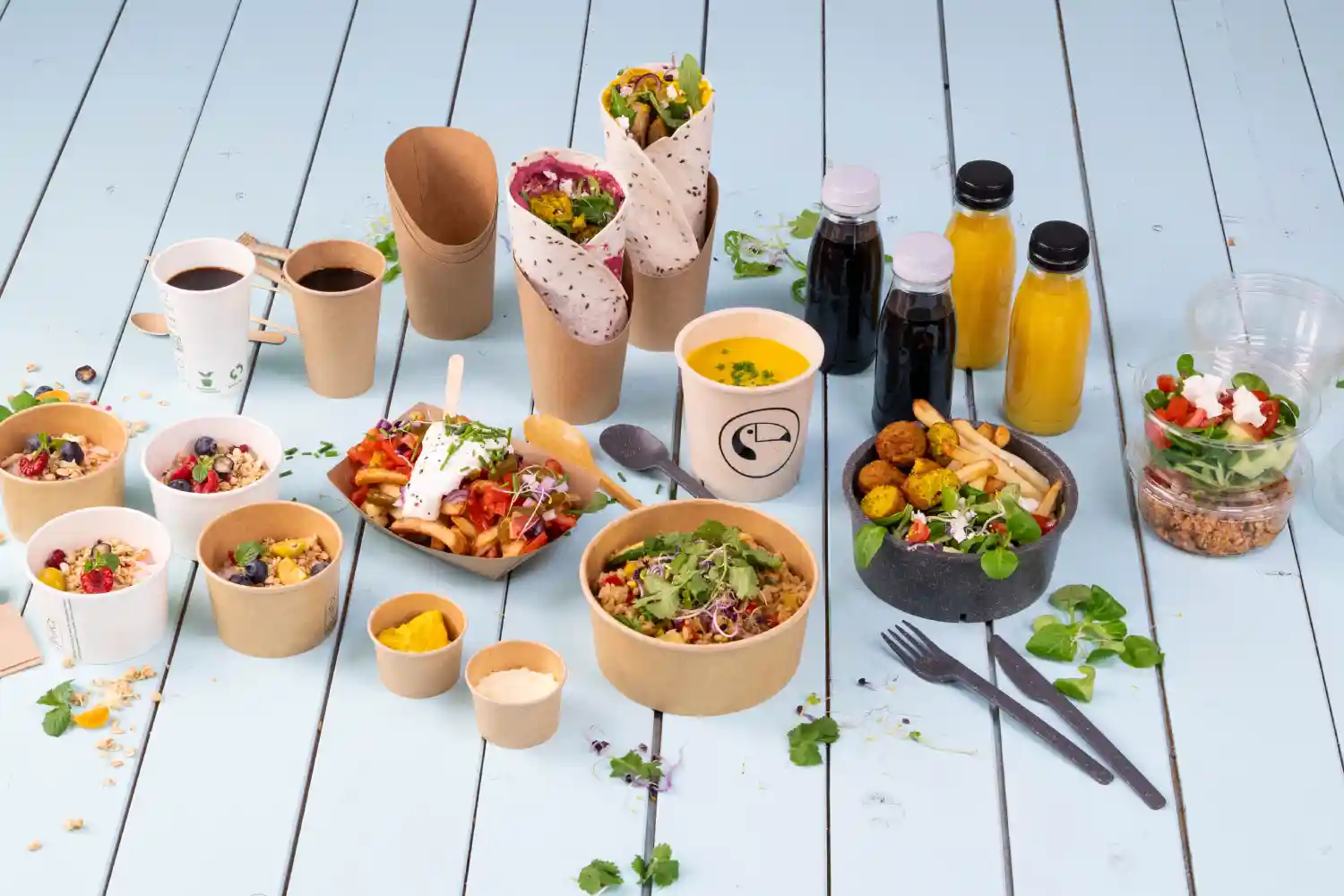 Assorted food items on a light blue wooden surface, including bowls of salad, wraps, drinks, and dessert options.