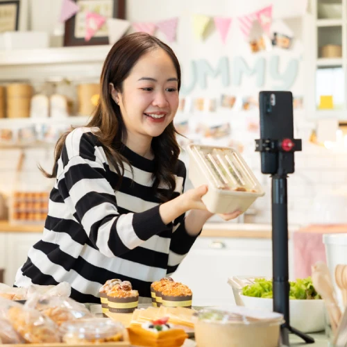 Woman in a striped sweater holds a food container while filming with a smartphone, surrounded by baked goods.