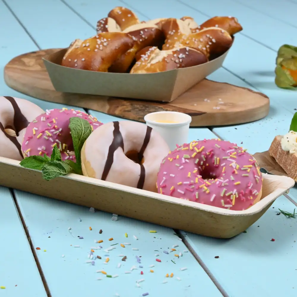 Three colorful donuts in a paper tray; two pink with sprinkles, one with chocolate drizzle. Pretzels are in the background.