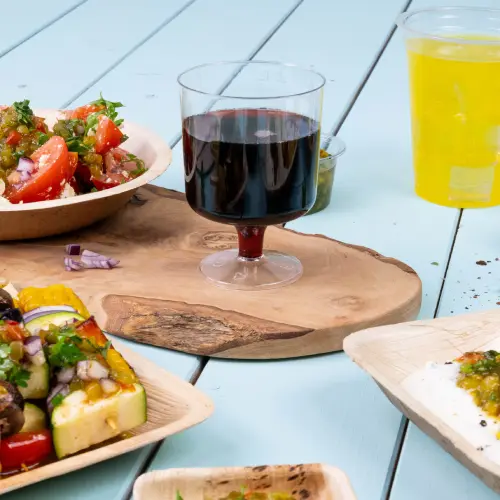 A clear glass cup with red liquid sits on a wooden board, surrounded by bowls of salad and grilled vegetables.