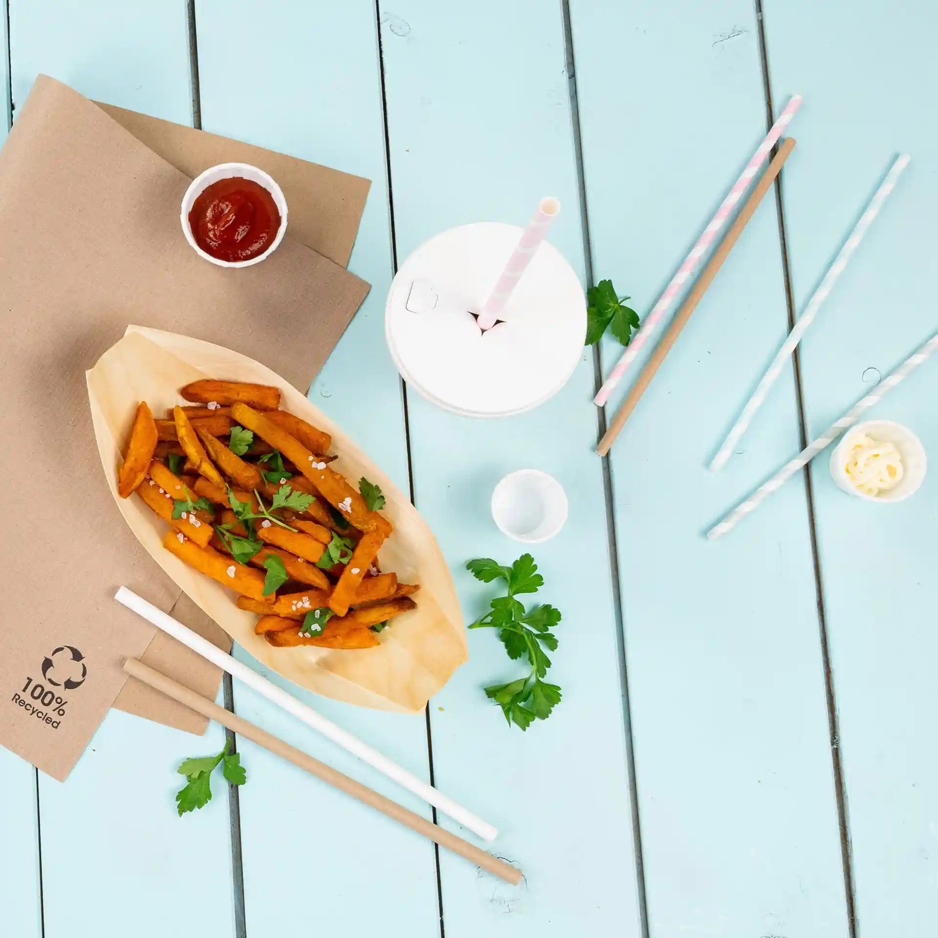 Image of sweet potato fries in a wooden boat with parsley, ketchup, a cup with straws, and paper straws on a blue surface.