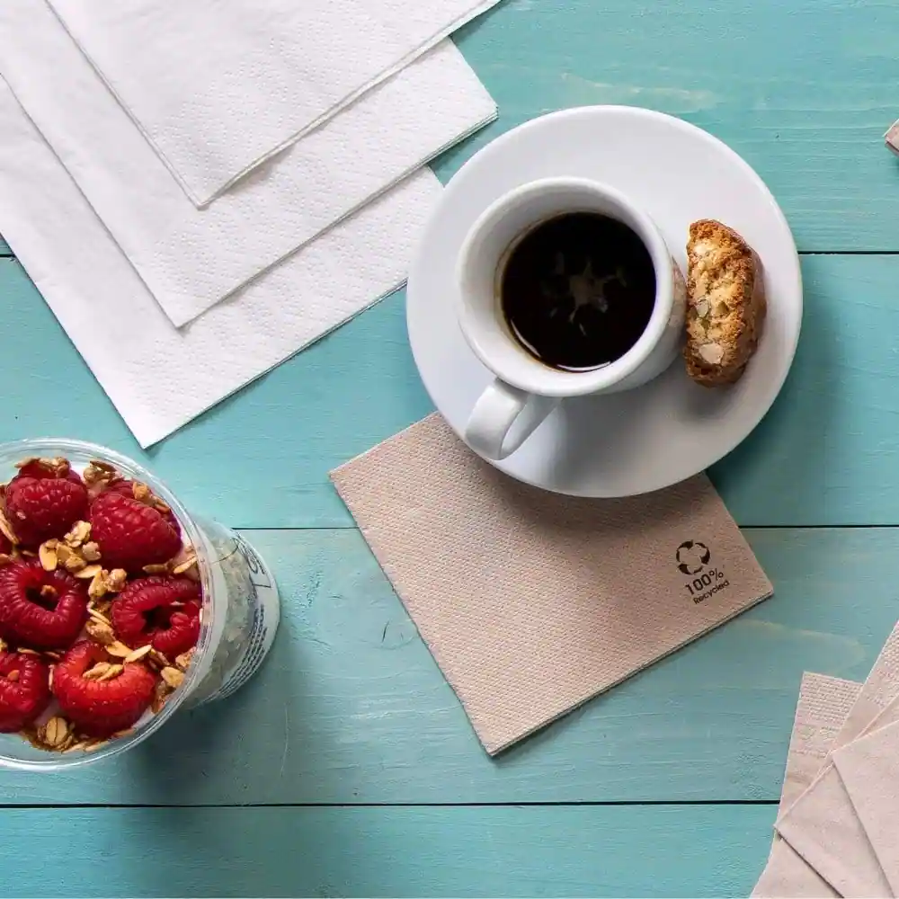 Cup of coffee on a saucer with a biscotti, next to a clear container of raspberries and granola, on a blue wooden surface.