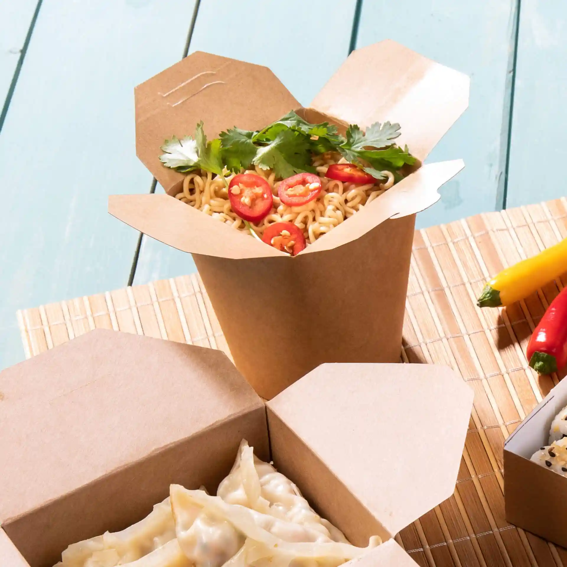 Brown cardboard noodle box filled with noodles, topped with sliced chili and cilantro, on a bamboo mat.