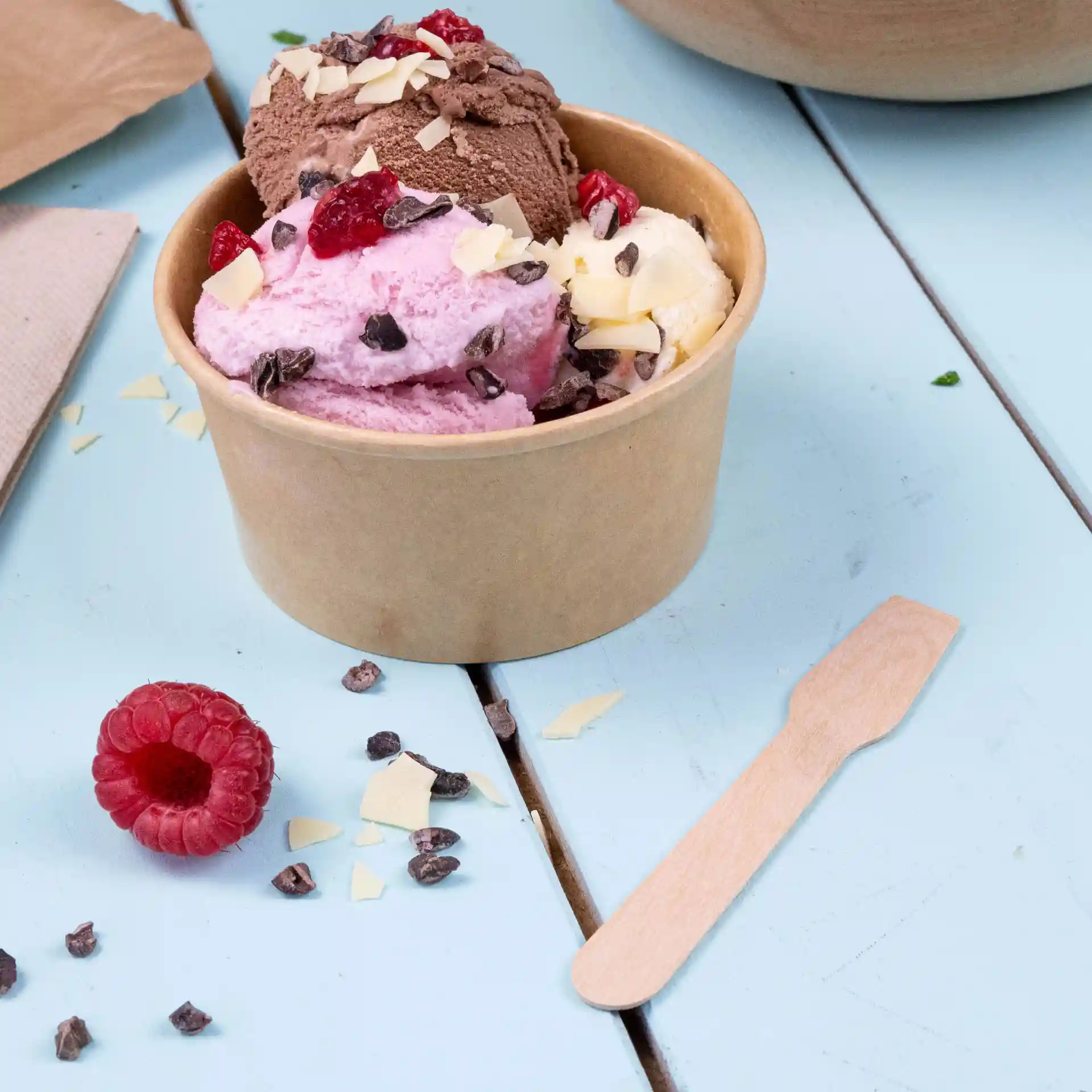 Cup containing three scoops of ice cream in pink, brown, and white, with toppings, beside a wooden spoon and a raspberry.