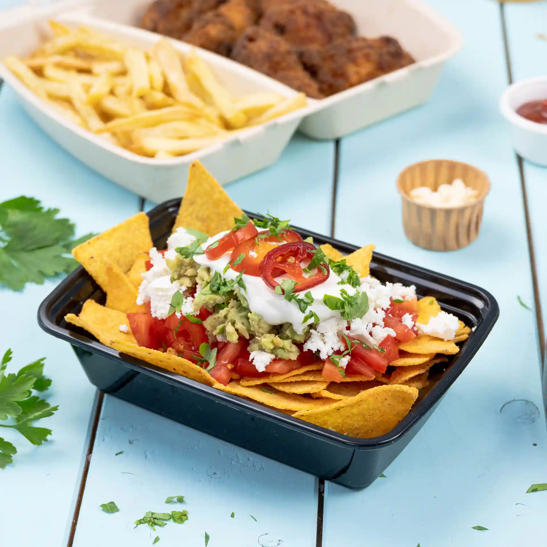 Black rectangular reusable container filled with nachos, topped with tomatoes, guacamole, sour cream, and cilantro.