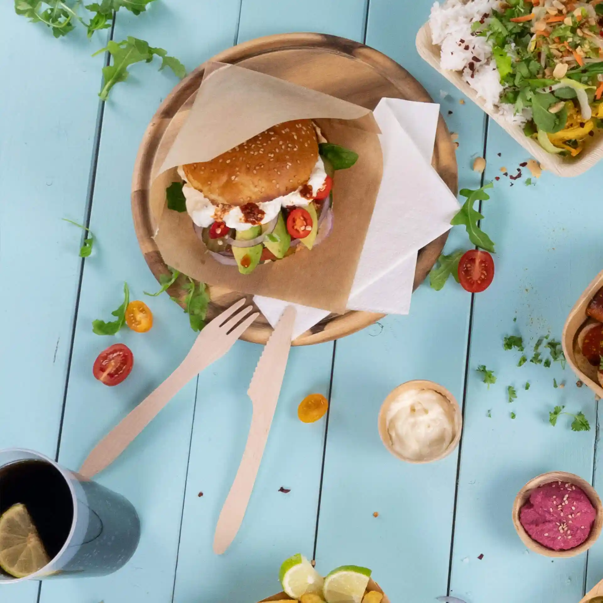 Image of a sesame seed bun sandwich with vegetables on a wooden plate, accompanied by wooden cutlery and a drink.