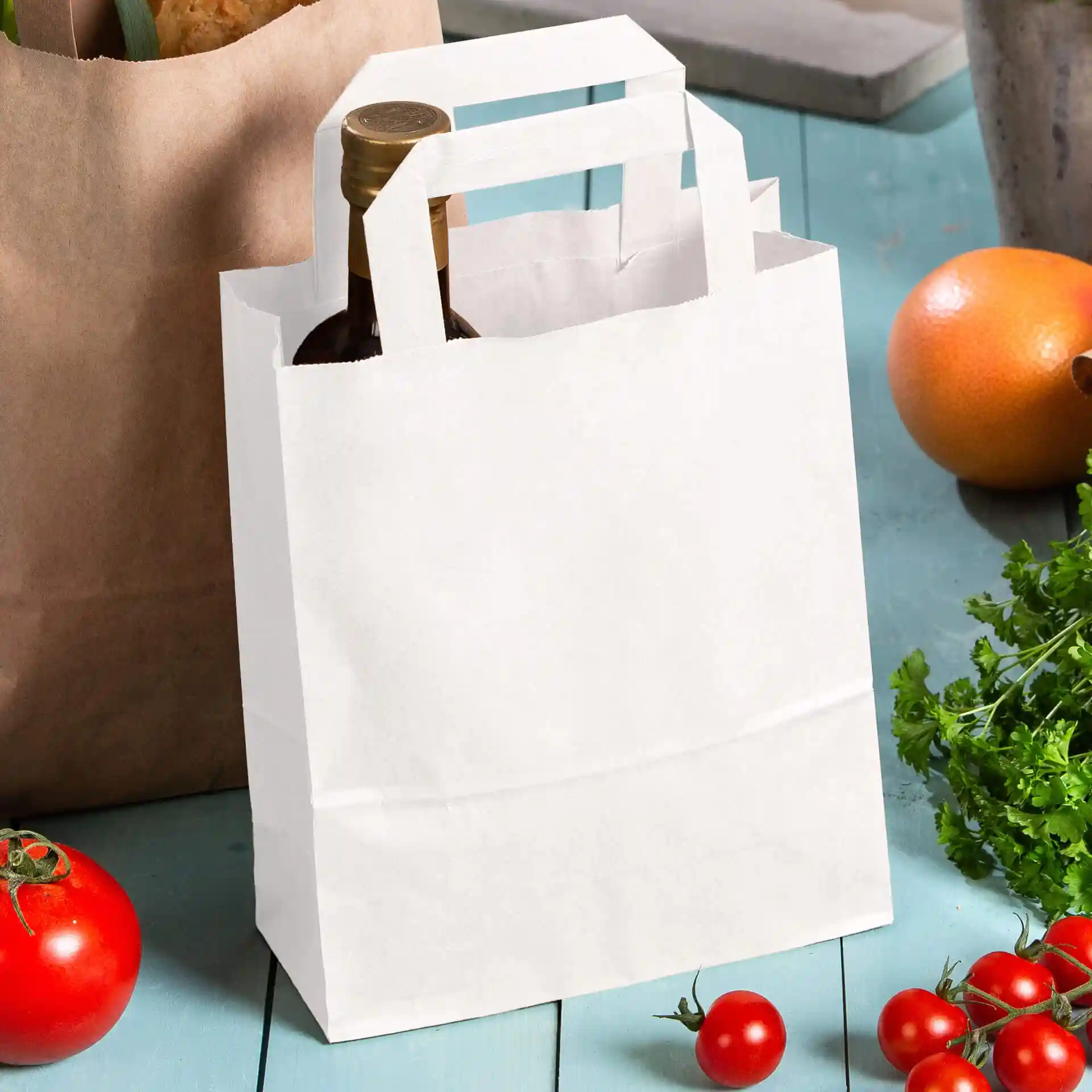 White kraft paper bag with handles, holding a bottle, surrounded by tomatoes and an orange on a blue surface.