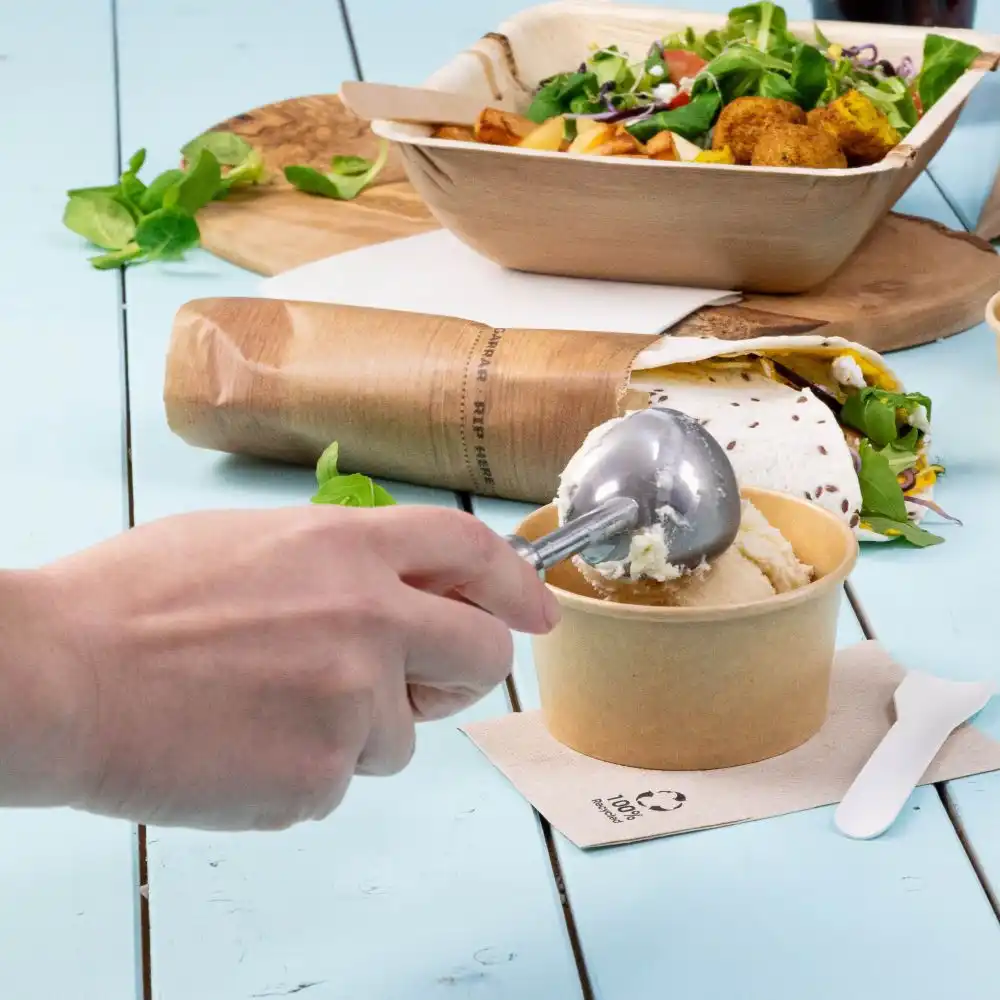 Hand holding a metal ice cream scoop over a paper cup of ice cream; salad and taco in eco-friendly containers nearby on a blue table.